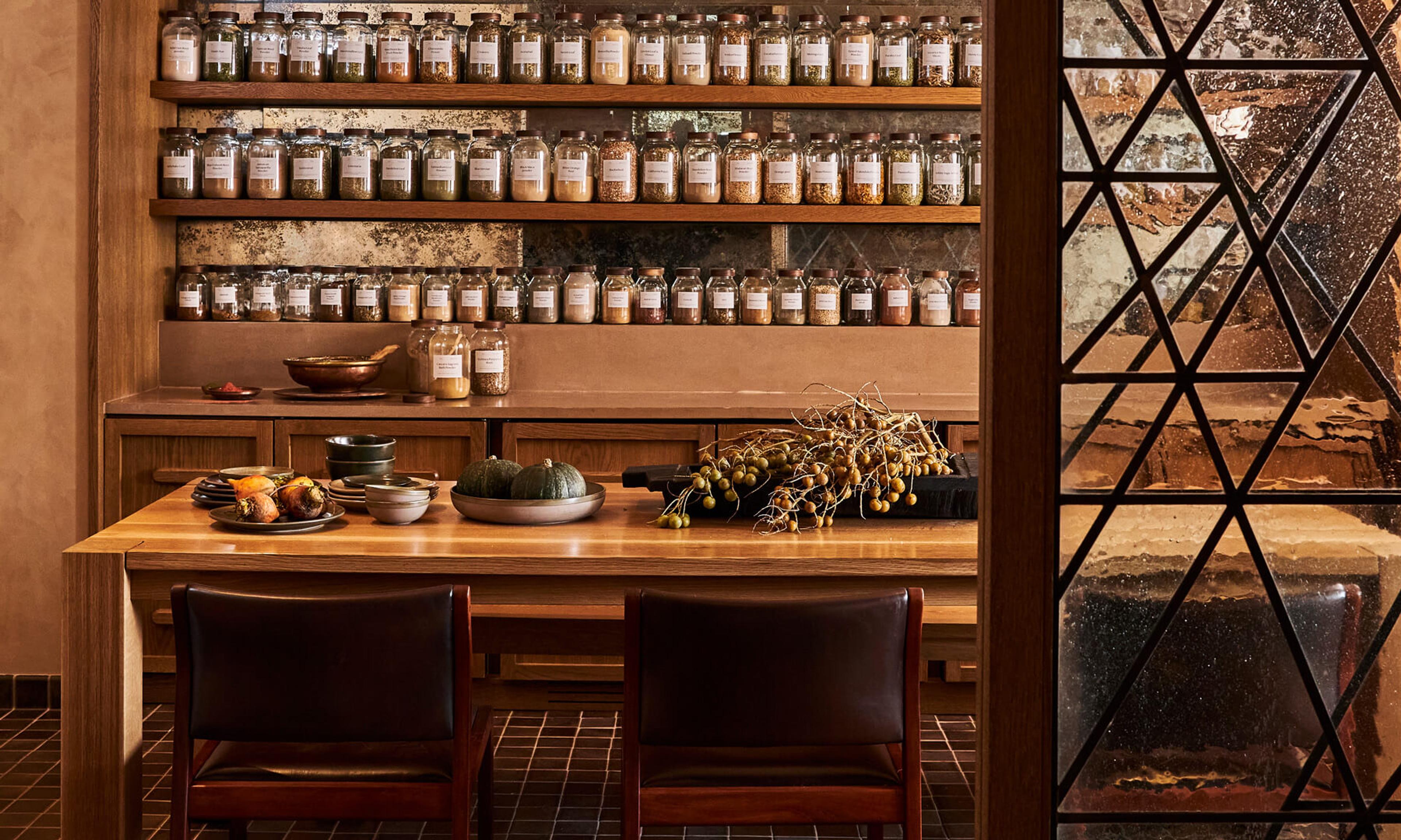 A wooden table set with bowls of food and small pumpkins in front of shelves lined with labelled spice jars.