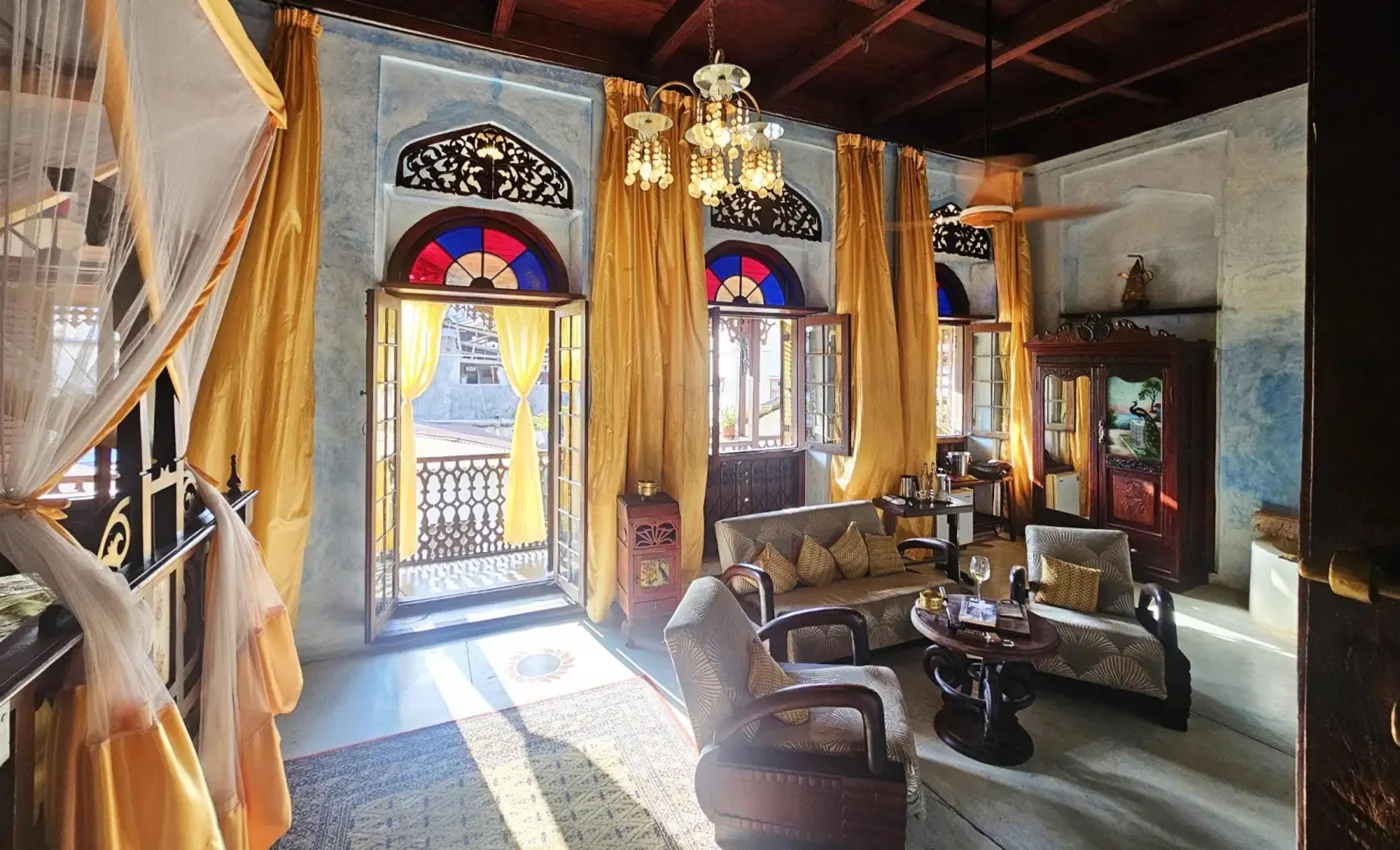 a high-ceilinged lounge with mustard silk curtains framing arched stained-glass windows open to a wrought-iron balcony, furnished with antique sofas and armchairs