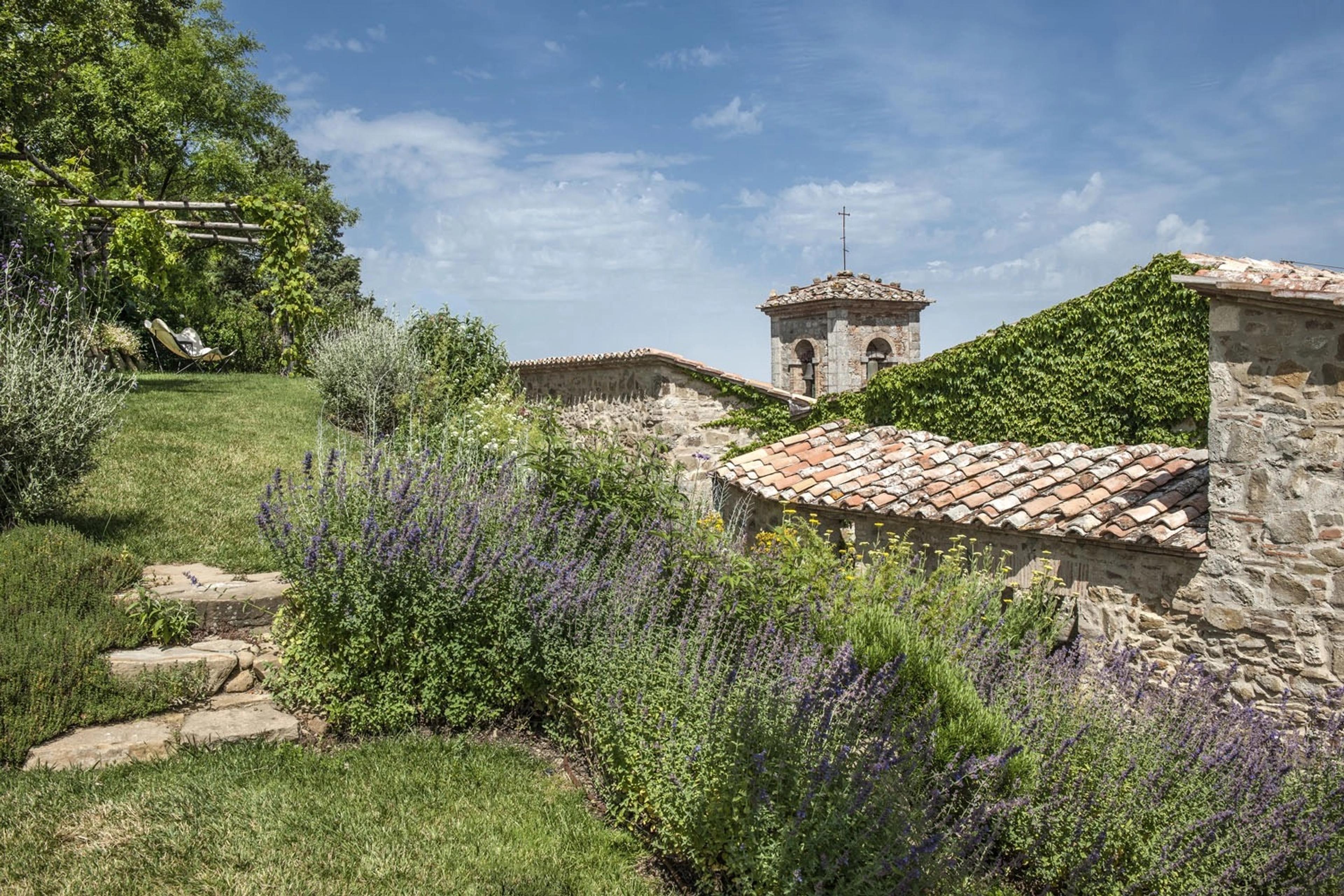 A garden path edged with lavender leading to a stone building with a bell tower, a sun lounger visible in the distance.