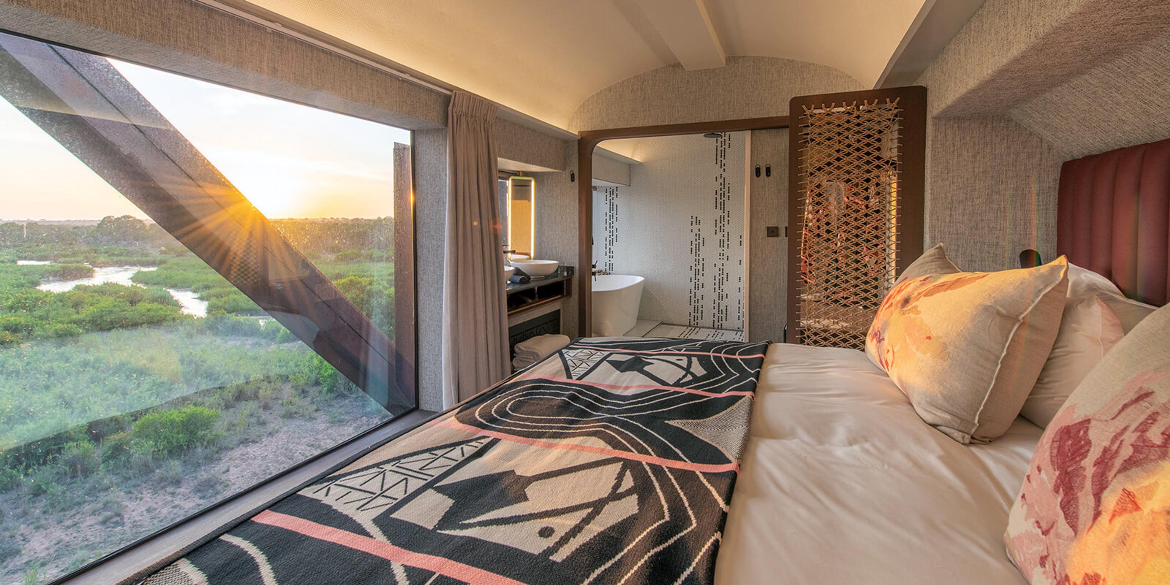 A bed inside a train carriage room facing a floor-to-ceiling window over wetland scrub at sunrise, with a freestanding bathtub visible through a screen beside it.