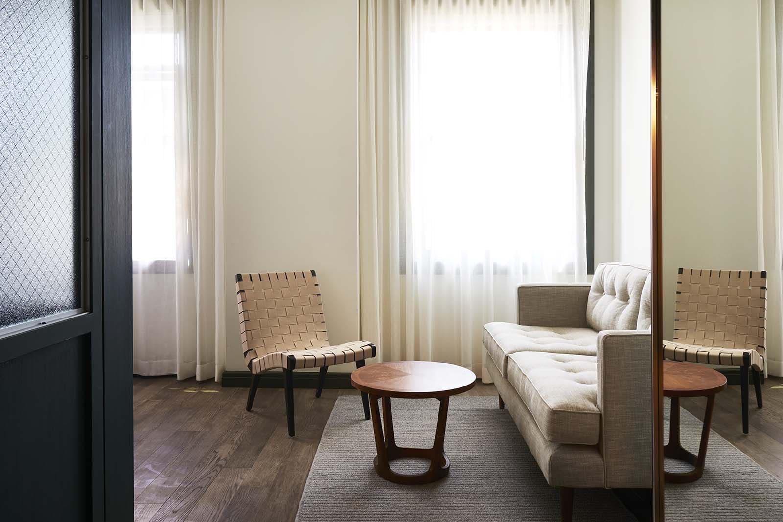 a beige sofa and a woven-strap armchair around a round wood coffee table on a grey rug, facing two curtained windows, reflected in a full-length mirror