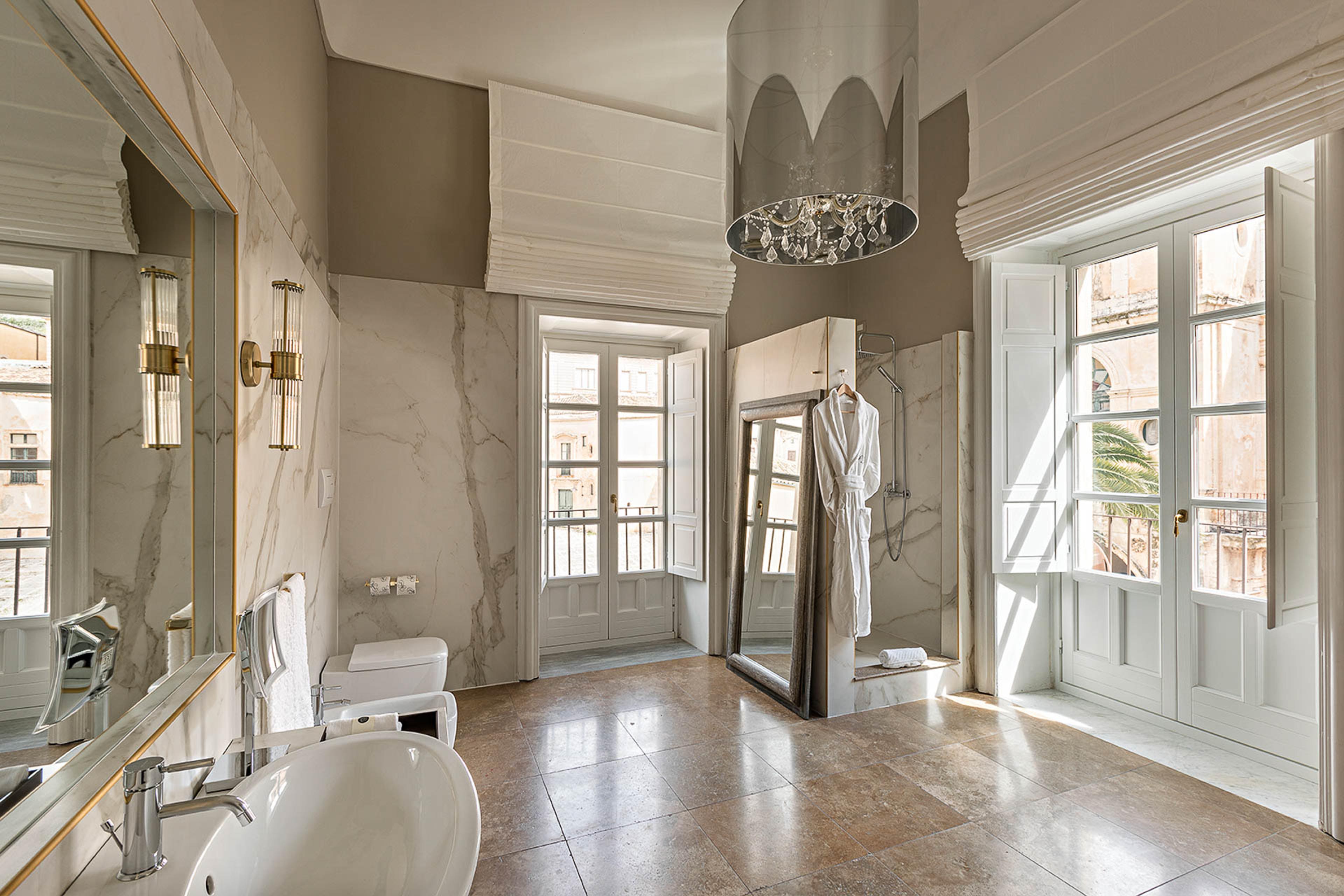 A marble bathroom with a freestanding mirror beside an open shower, a bathrobe hanging on a hook, and two sets of French doors open to a street of stone buildings.