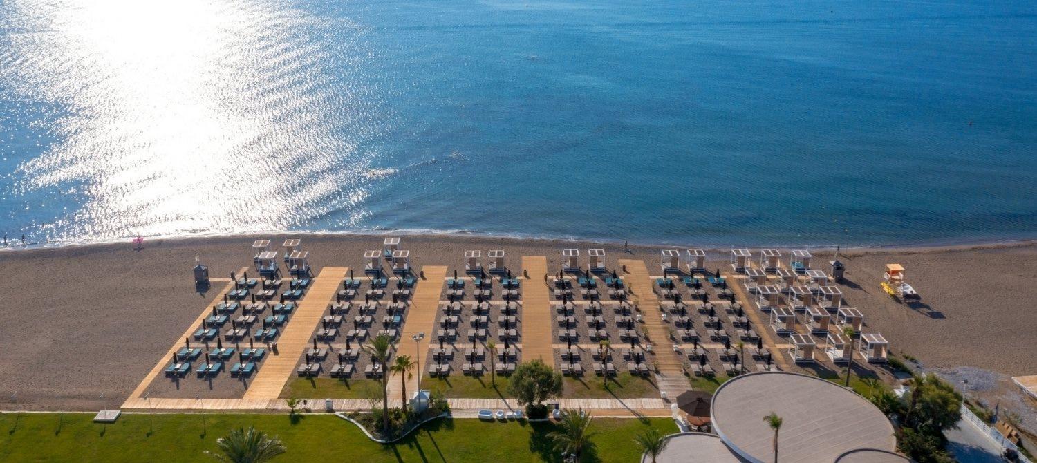 An aerial view of rows of beach sunbeds and shade canopies on sand beside the sea.