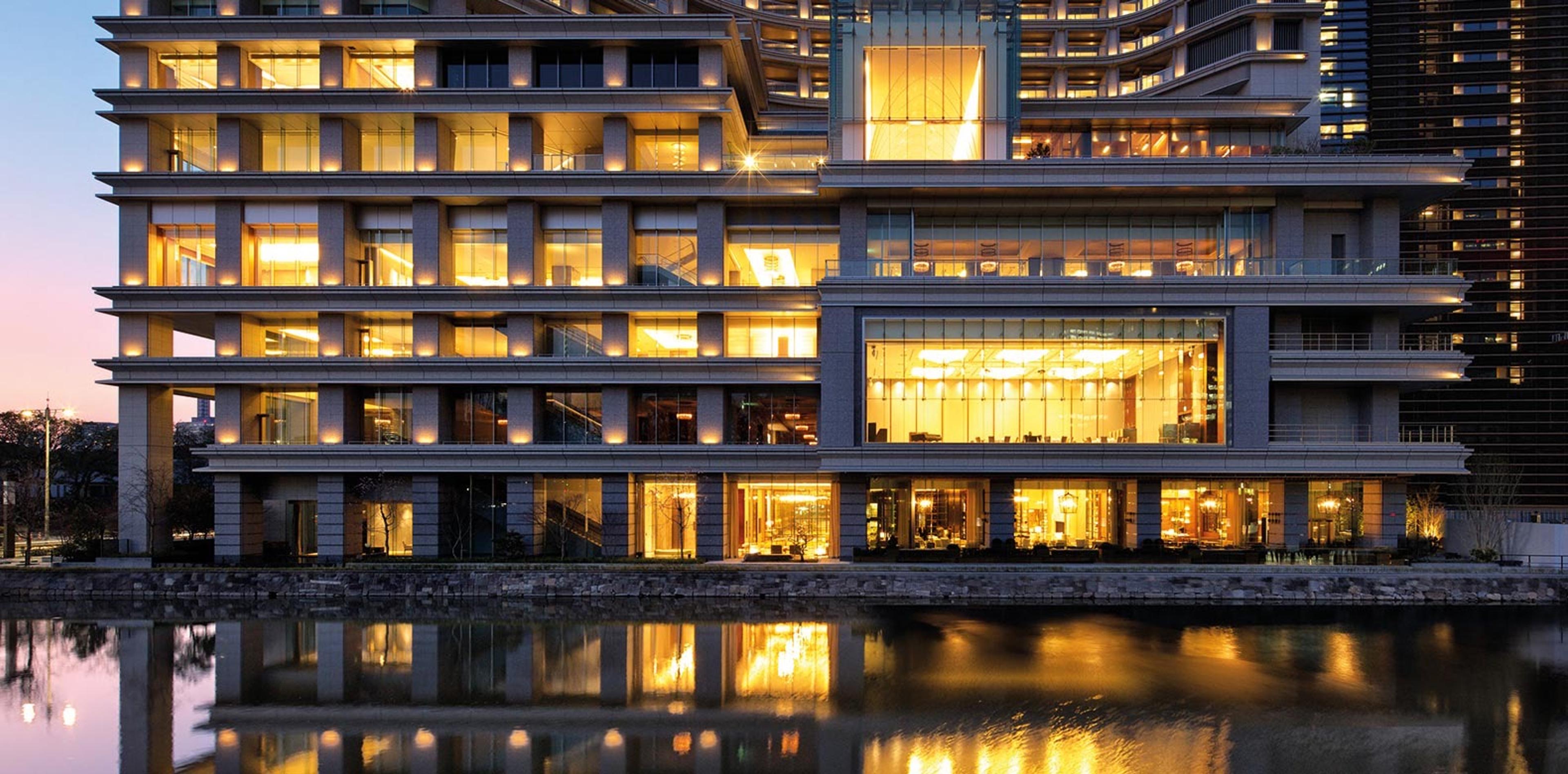 A hotel tower lit from within at dusk, its glass front reflected in the water in front of it.