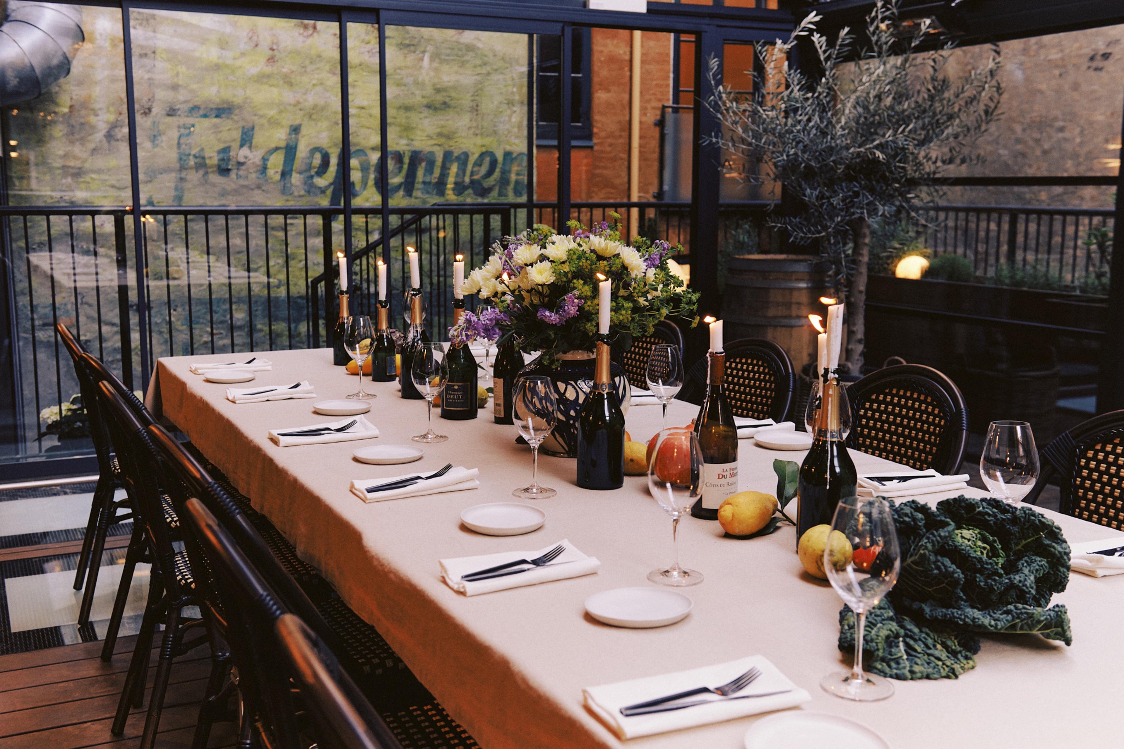 A long table set for dinner outdoors with candles, flowers, wine bottles and fruit, next to glass doors and a potted tree, at dusk.