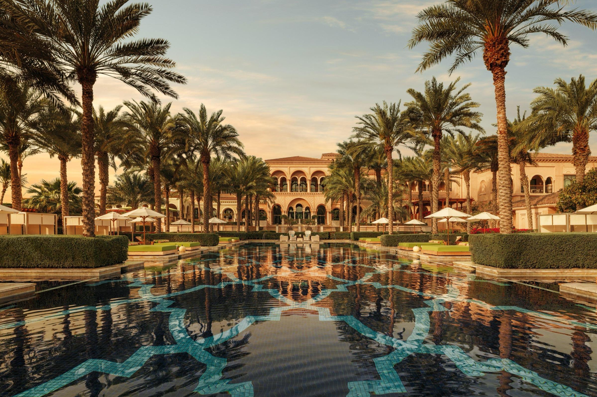 A long reflecting pool with a blue mosaic pattern leads between rows of palm trees to a two-story building with arched windows, lit at dusk.