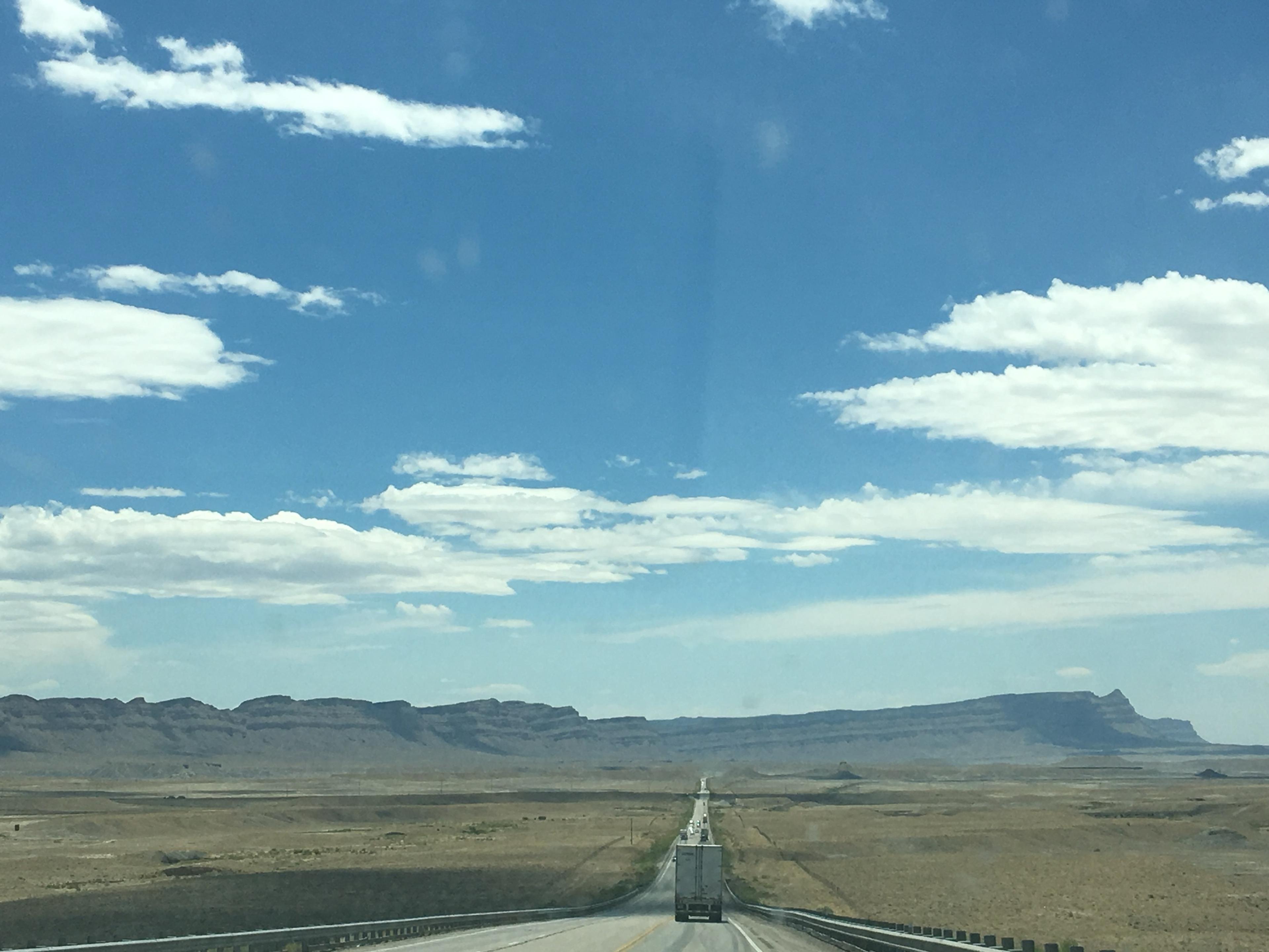 A straight, empty highway running across scrubland towards a distant flat-topped mesa, with white clouds overhead