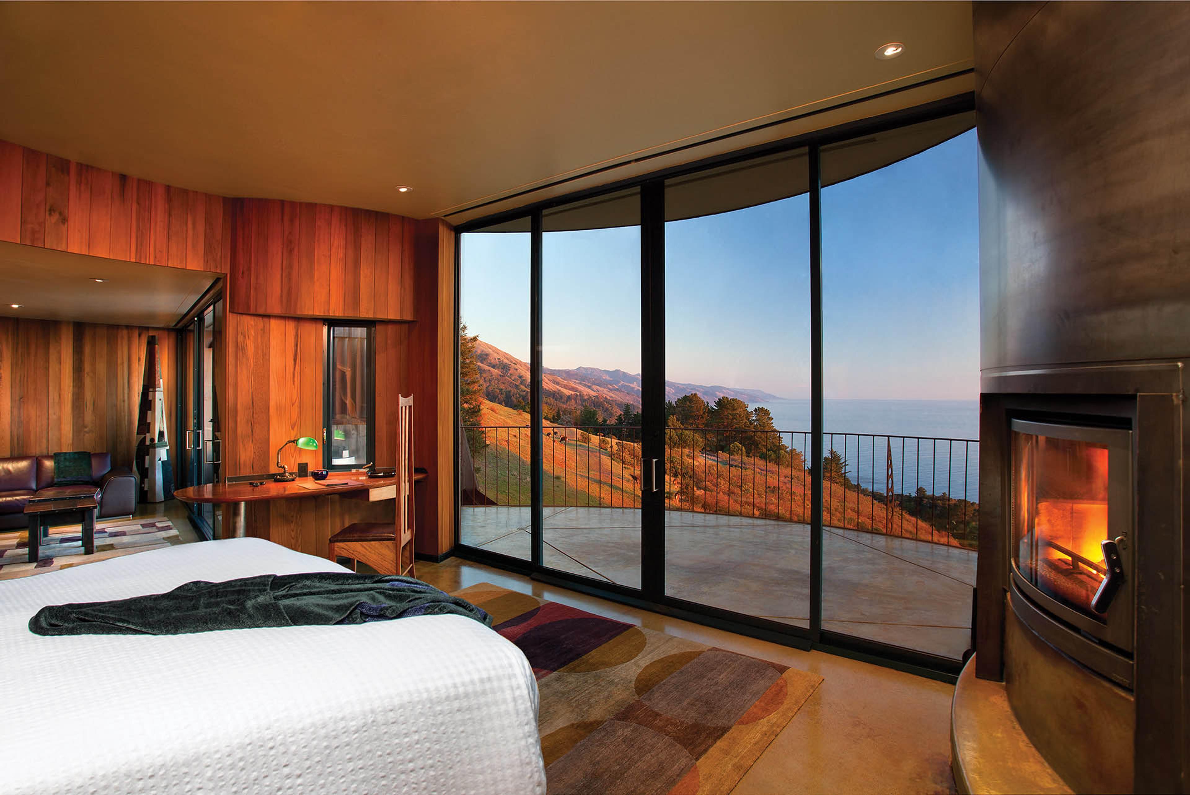 A bedroom with a white-made bed and a wood desk and chair, a lit wood-burning stove to the right, and a wall of glass doors open onto a terrace overlooking hillside and ocean at dusk.