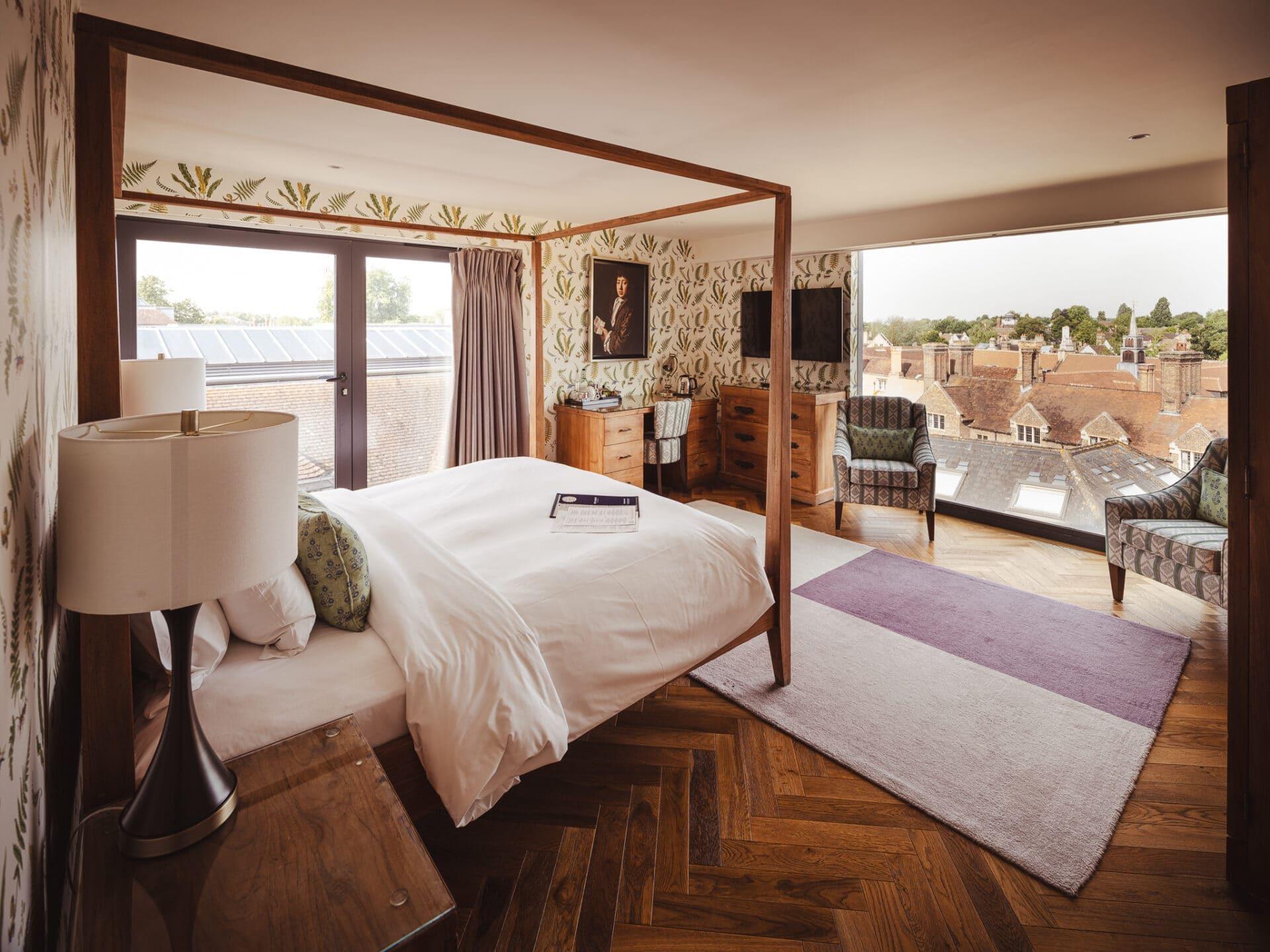 a dark wood four poster bed in a hotel suite with botanical wallpaper, a framed portrait, and a view over Cambridge rooftops through floor to ceiling windows