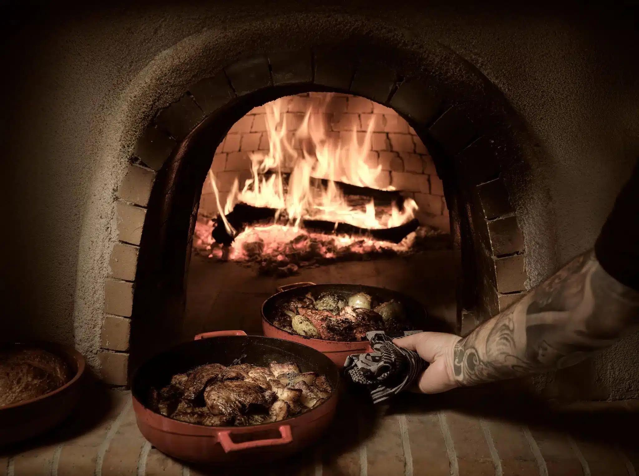 Two copper pans of roasted meat and potatoes being lifted from a blazing wood-fired oven by a tattooed arm.