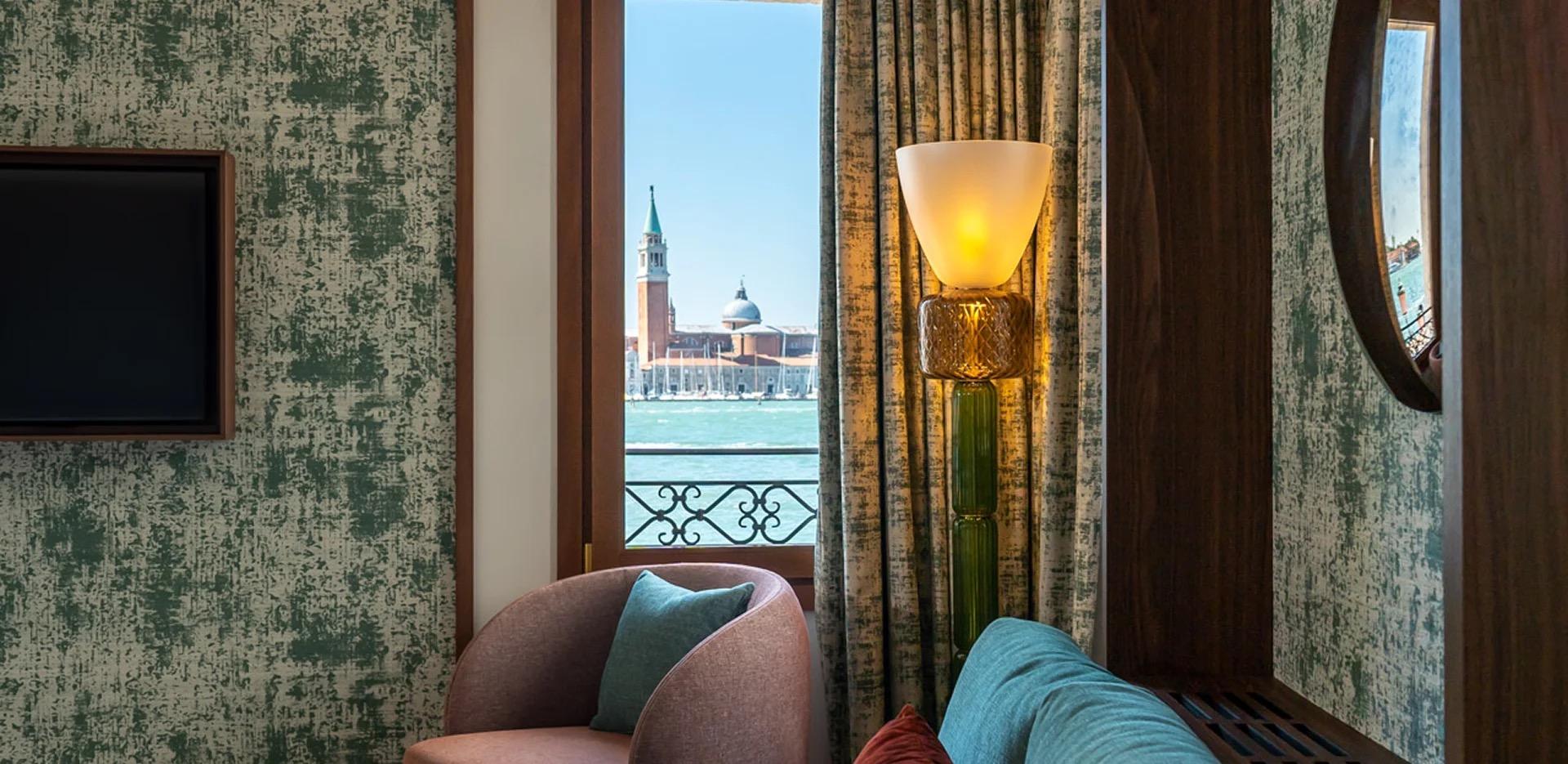 An armchair and sofa beside a curtained window looking across water to a domed church and bell tower.