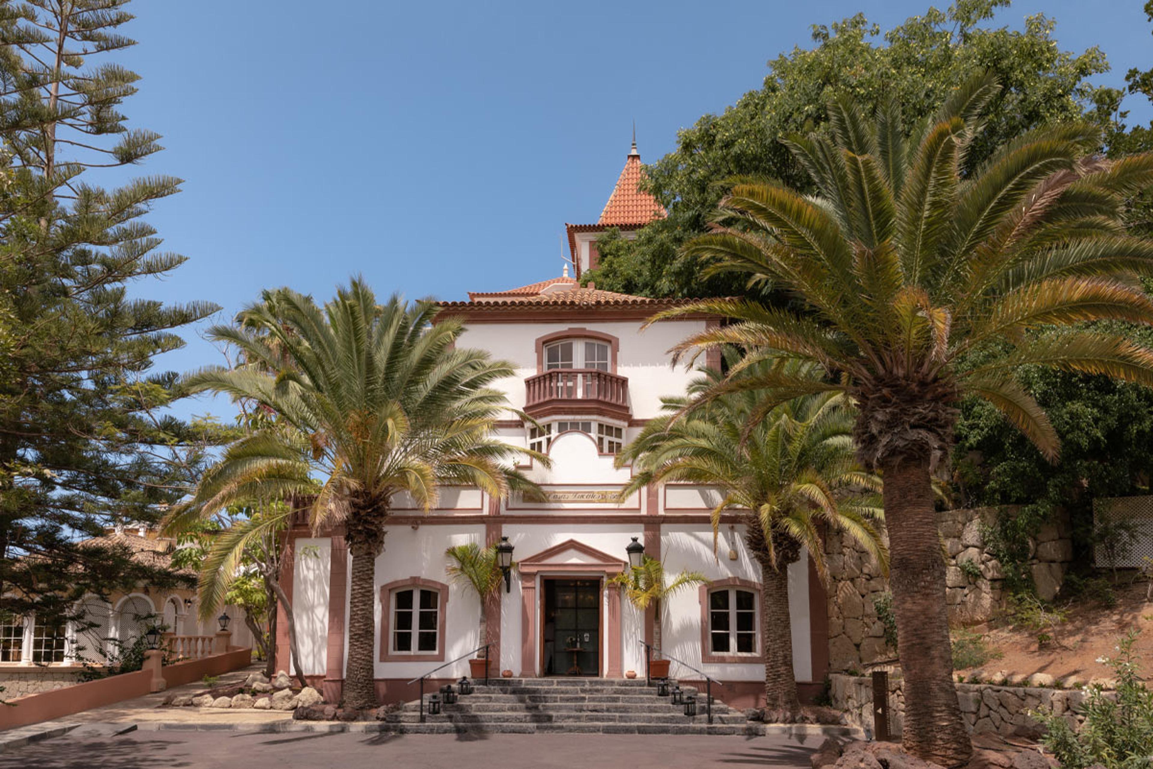 A colonial-style white and terracotta building with a corner turret, approached by steps and framed by palm trees.