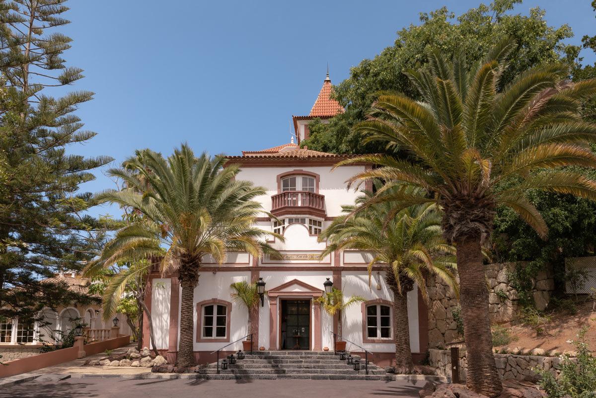 A colonial-style white and terracotta building with a corner turret, approached by steps and framed by palm trees.
