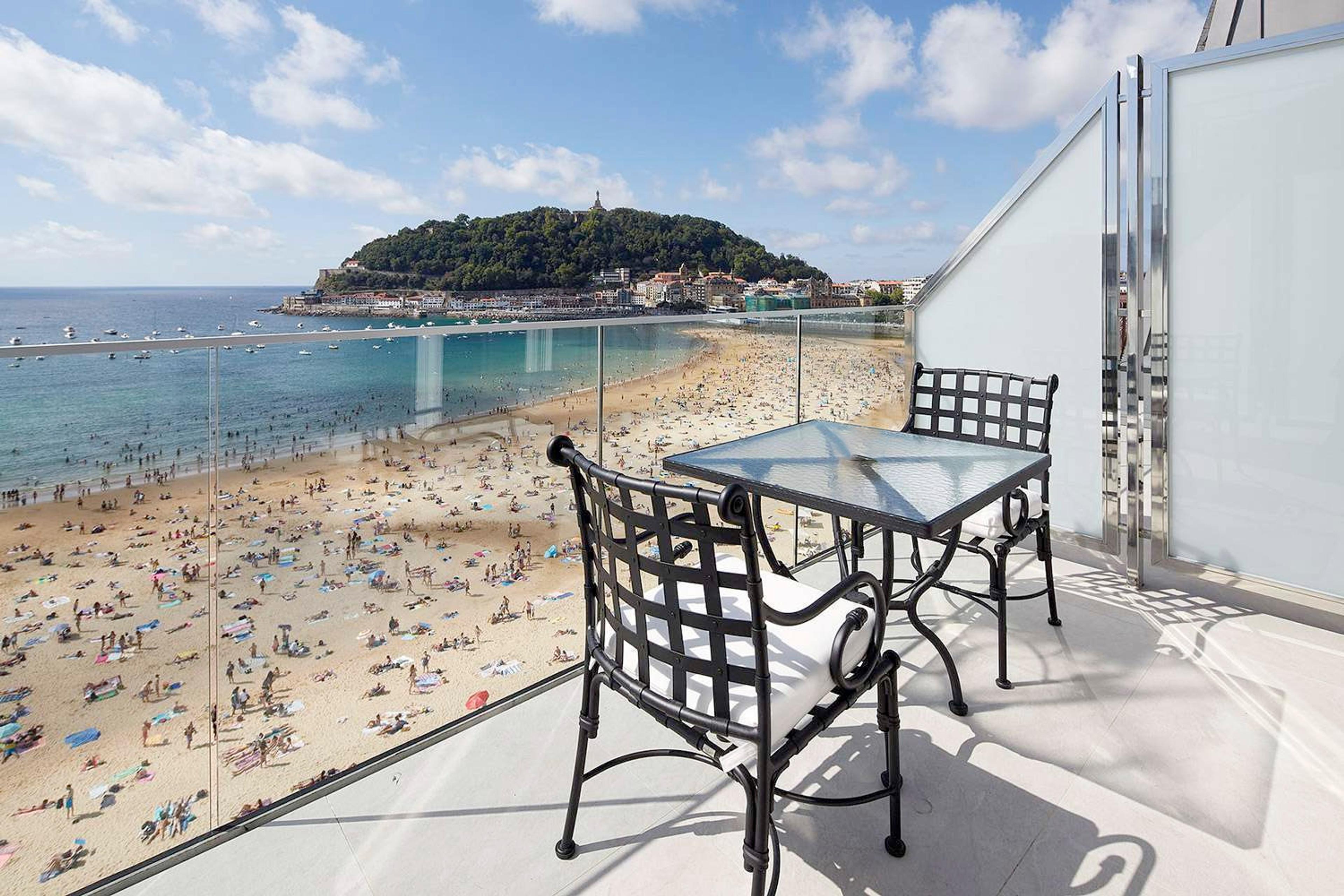 A balcony table and two wrought-iron chairs looking out over a crowded sandy beach and a tree-covered headland.