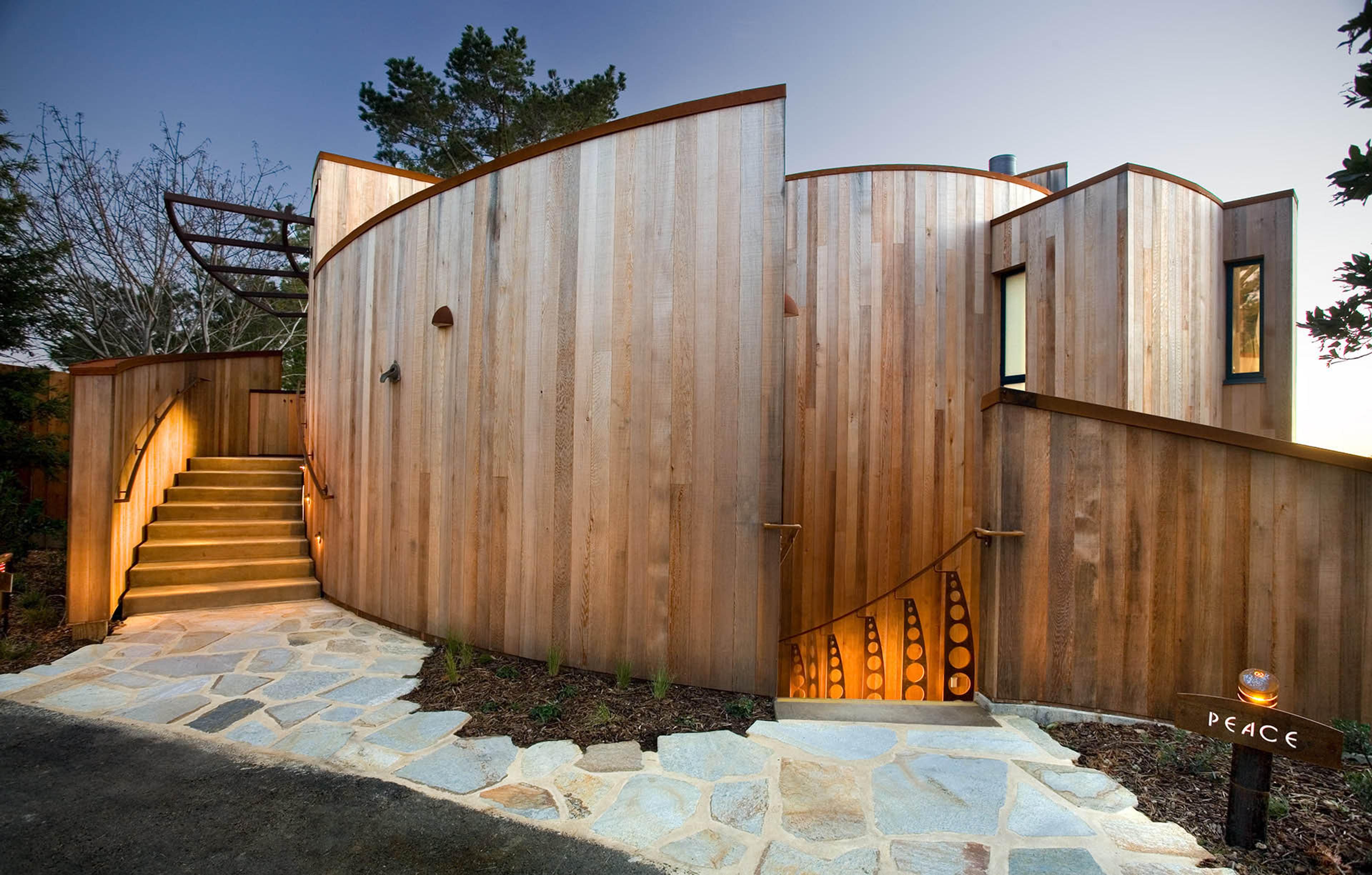 The exterior of a guest house at dusk, built from curved cedar paneling with copper trim, a lit stone stairway, and a small sign reading PEACE beside the entrance.