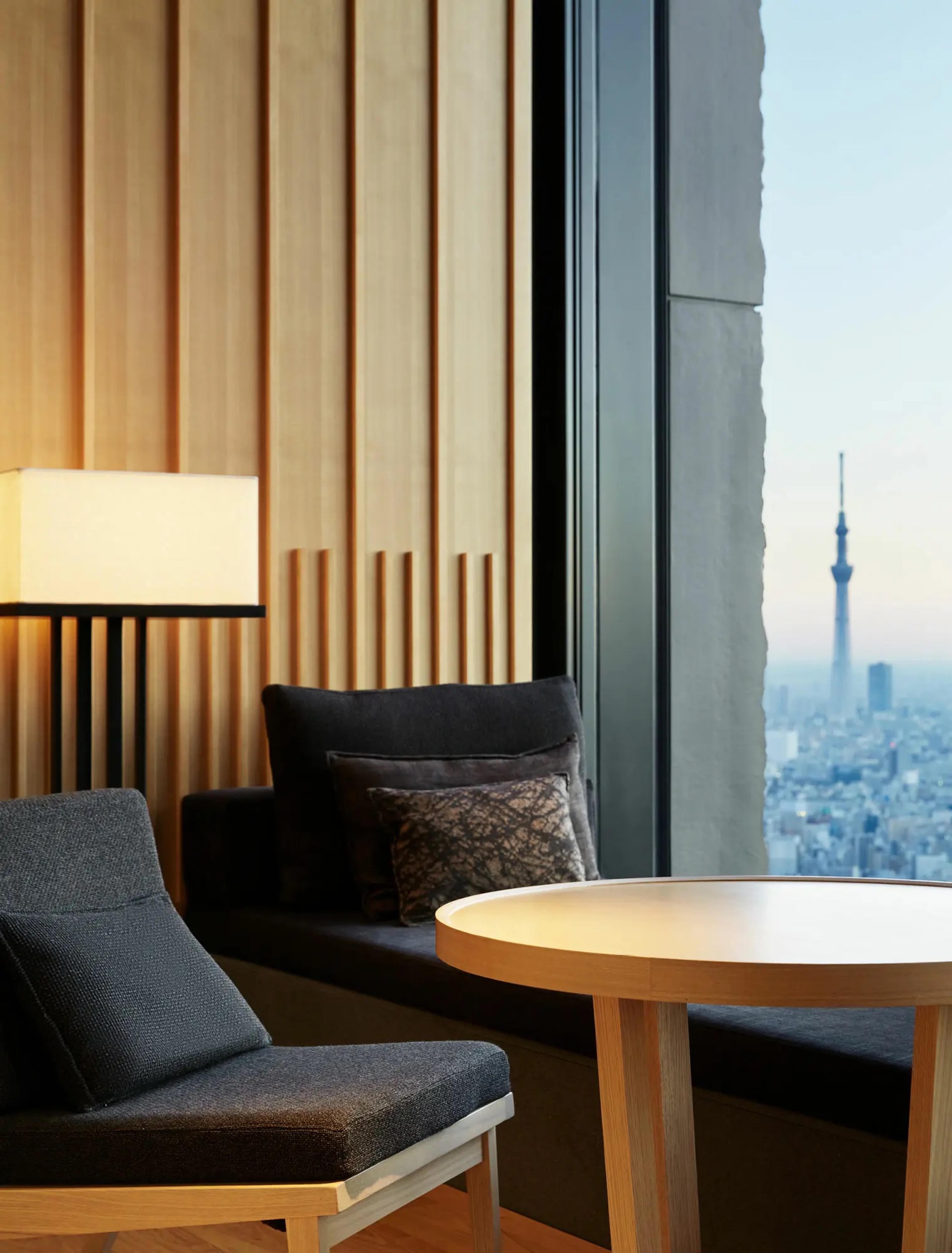 A window-side armchair and round table lit by a lamp, facing a floor-to-ceiling window with Tokyo Skytree on the skyline at dusk.