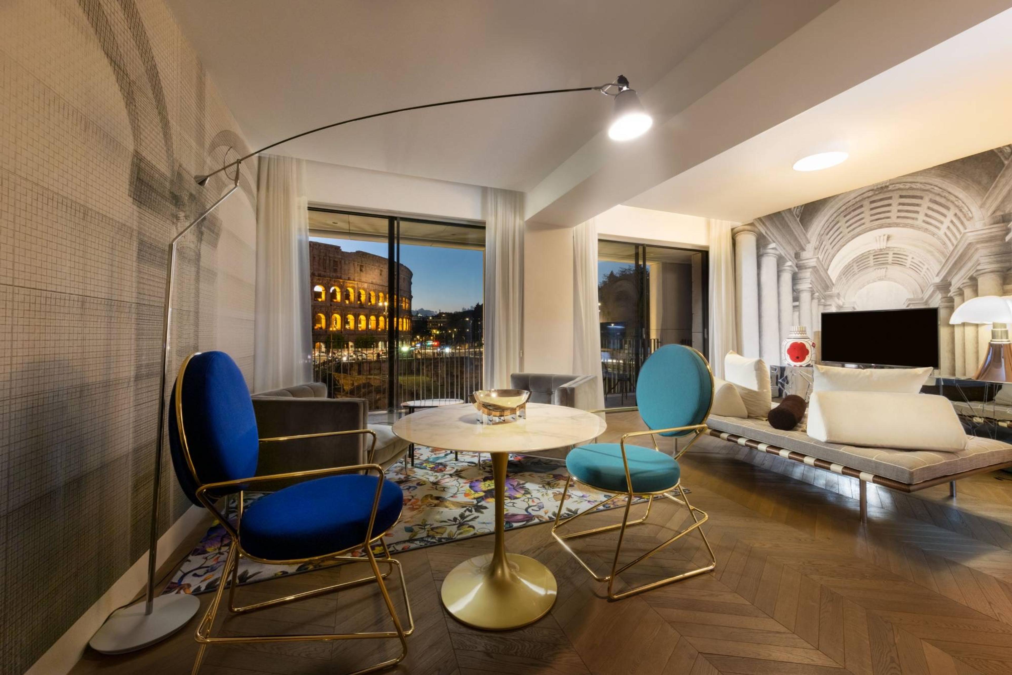 A living room at dusk with blue and teal chairs round a marble table, and the Colosseum lit through sliding glass doors, with a painted colonnade on the wall at right.