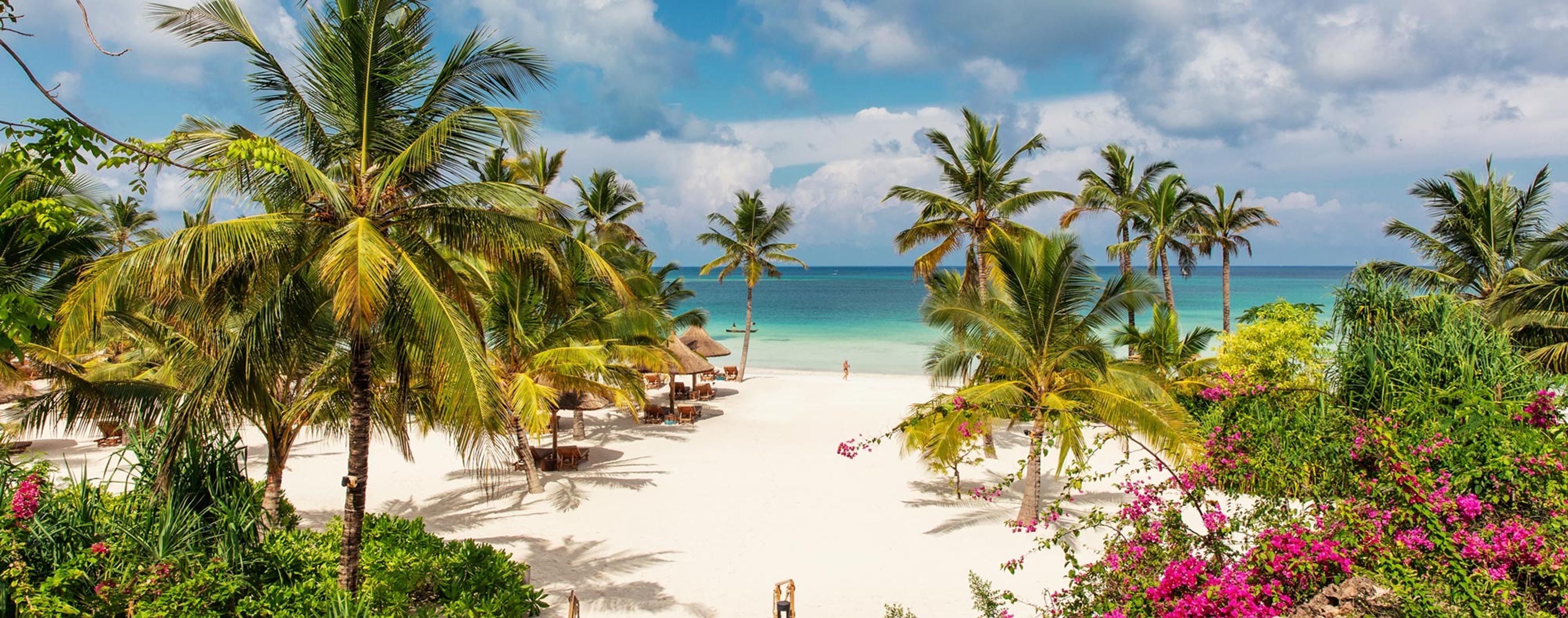 a white sand beach framed by palm trees and pink bougainvillea, with thatched sunshades and a person walking by the turquoise sea