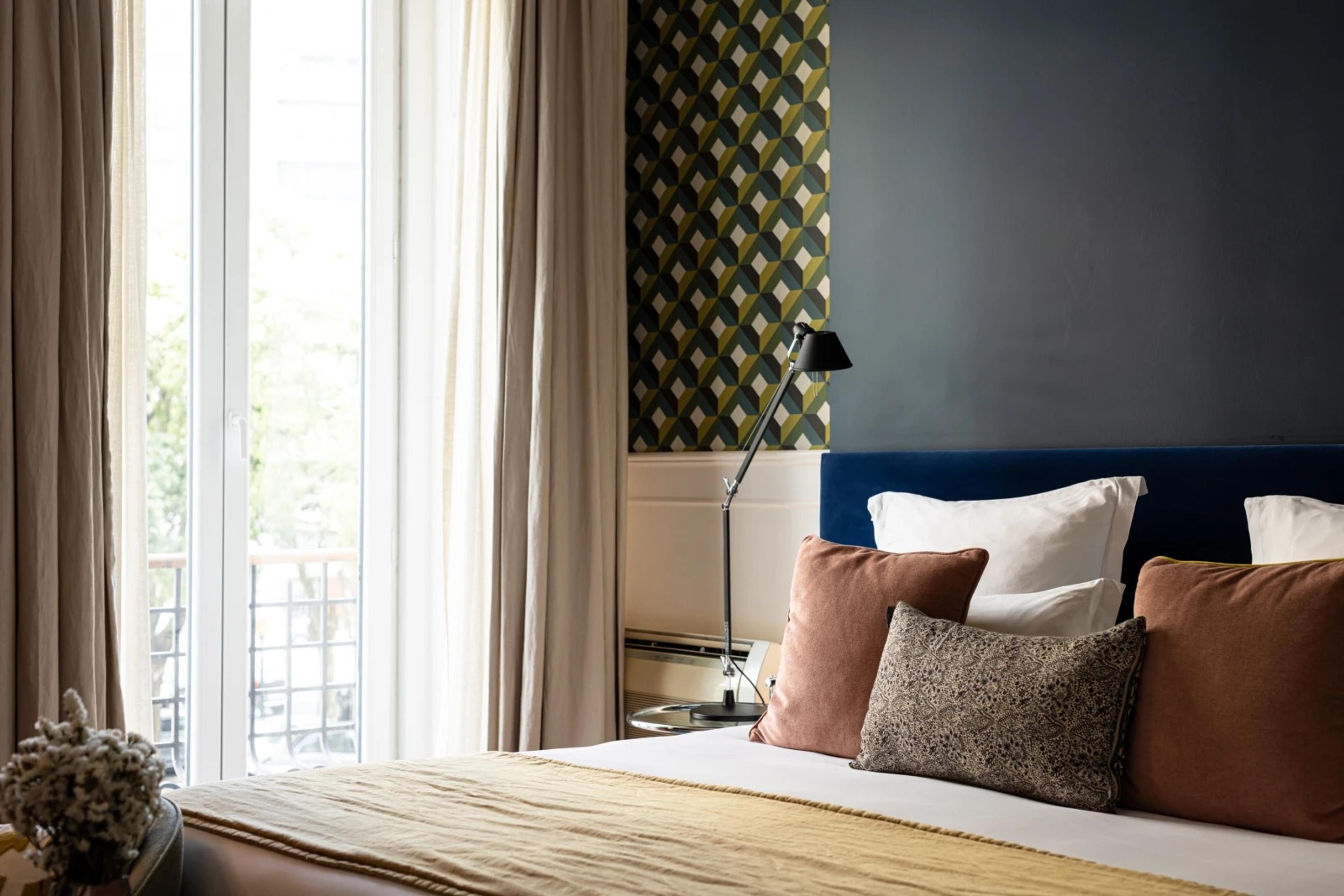 A made bed with a dark blue headboard and rust and tan cushions, beside a tall window with pale curtains open onto trees and a balcony, and a patterned wall with a black lamp.