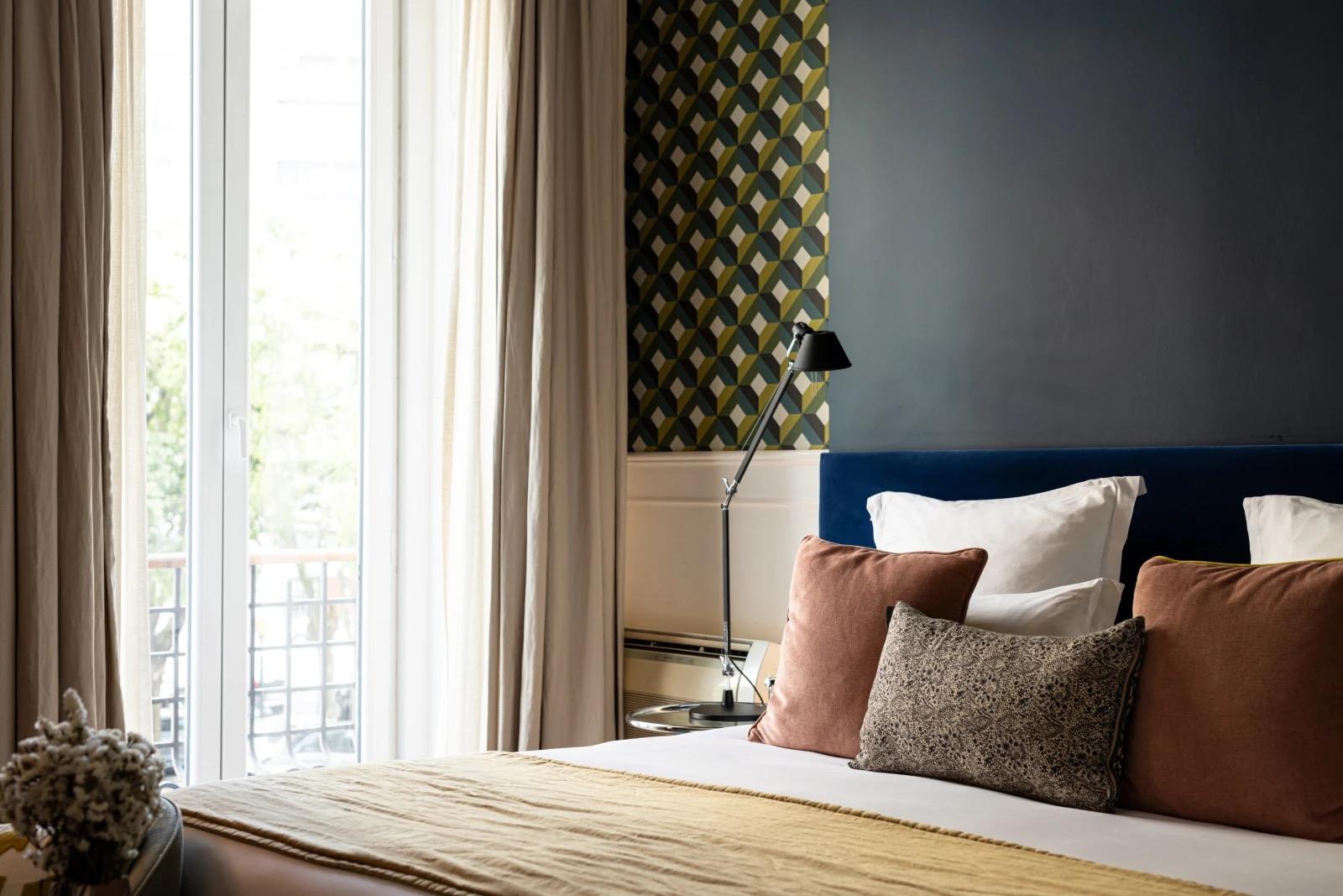 A made bed with a dark blue headboard and rust and tan cushions, beside a tall window with pale curtains open onto trees and a balcony, and a patterned wall with a black lamp.