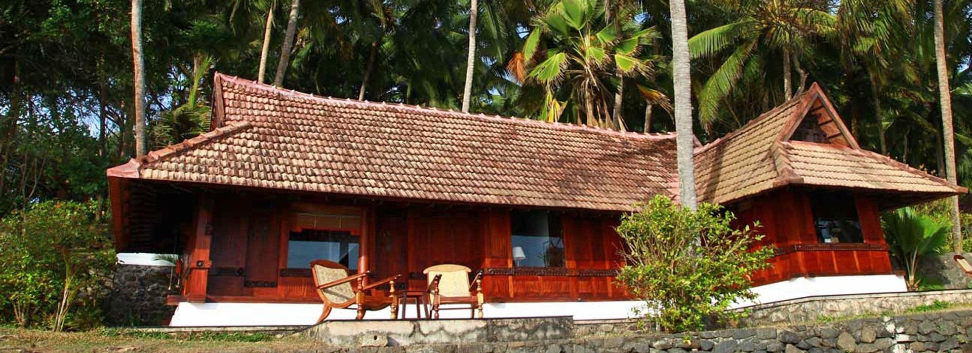 a long, low cottage with a red tiled roof and dark timber walls, set among palm trees under a clear blue sky