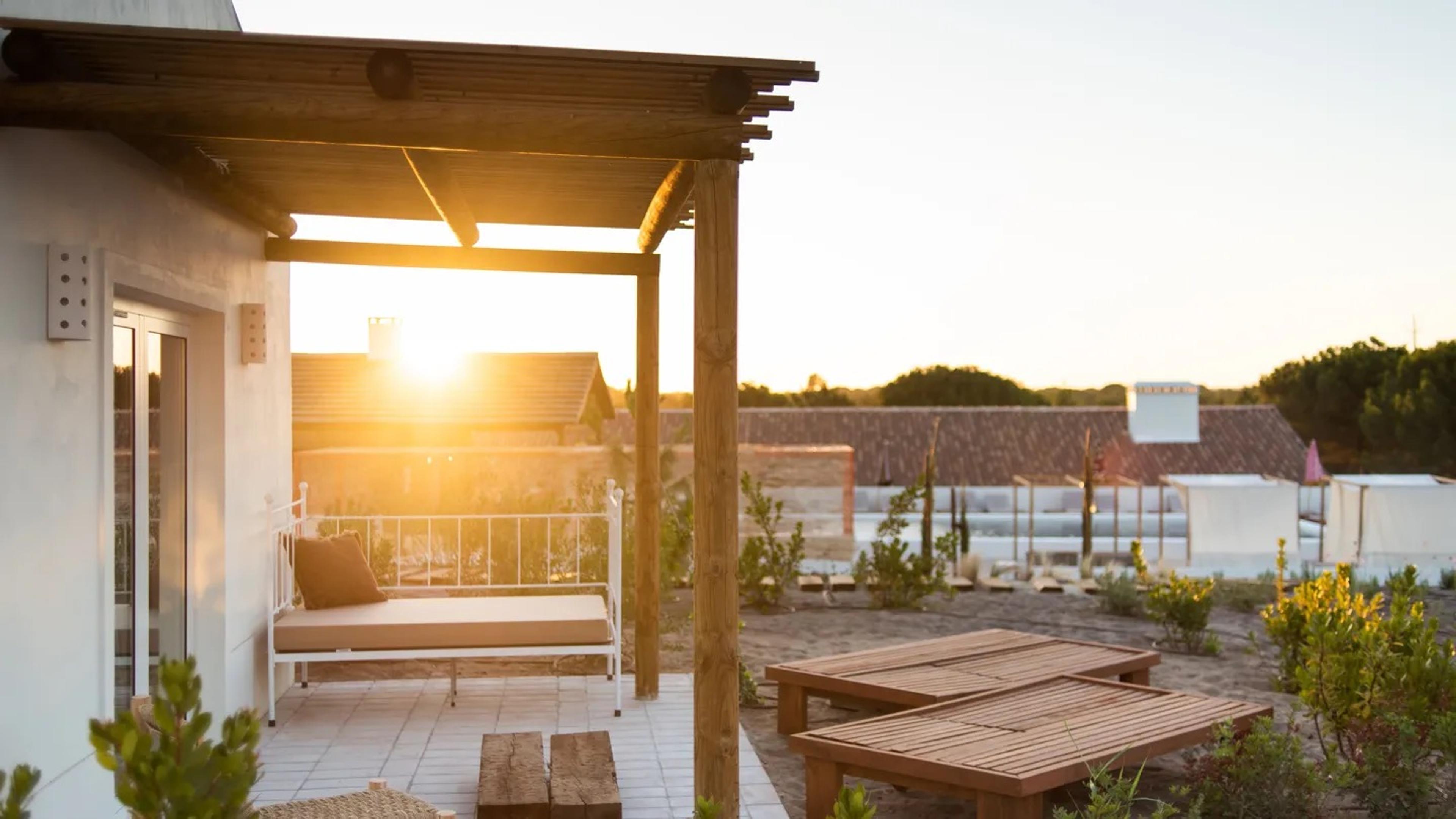 a covered terrace with a wooden pergola and a daybed beside two low benches, the sun setting behind rooftops with a pool visible in the distance