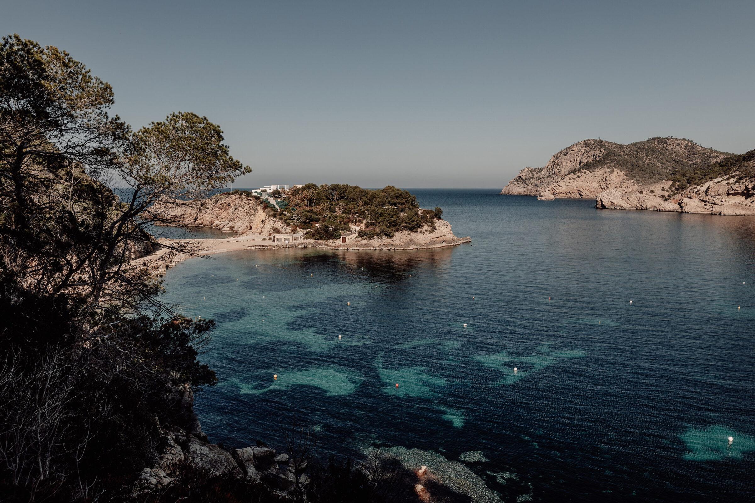 A small cove seen from a clifftop, with clear blue water, pines on a rocky point and a low white building on the headland across the bay.
