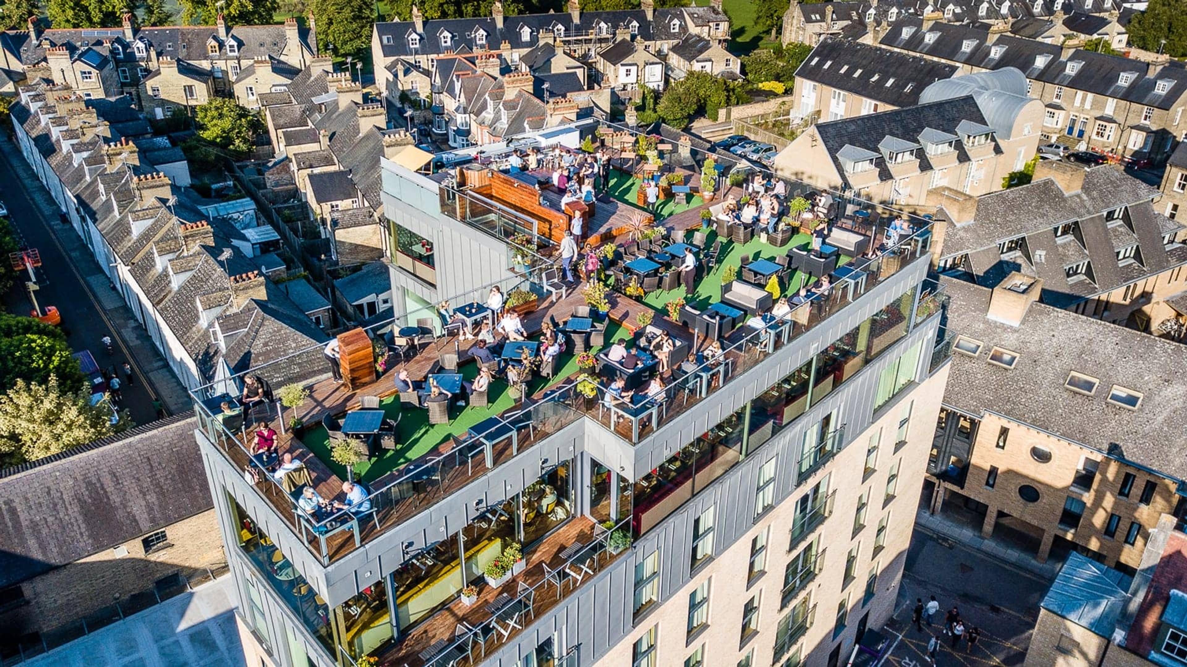 an aerial view at golden hour of the hotel's rooftop terrace and bar crowded with people, set above the gabled roofs of Cambridge with a church tower in the distance
