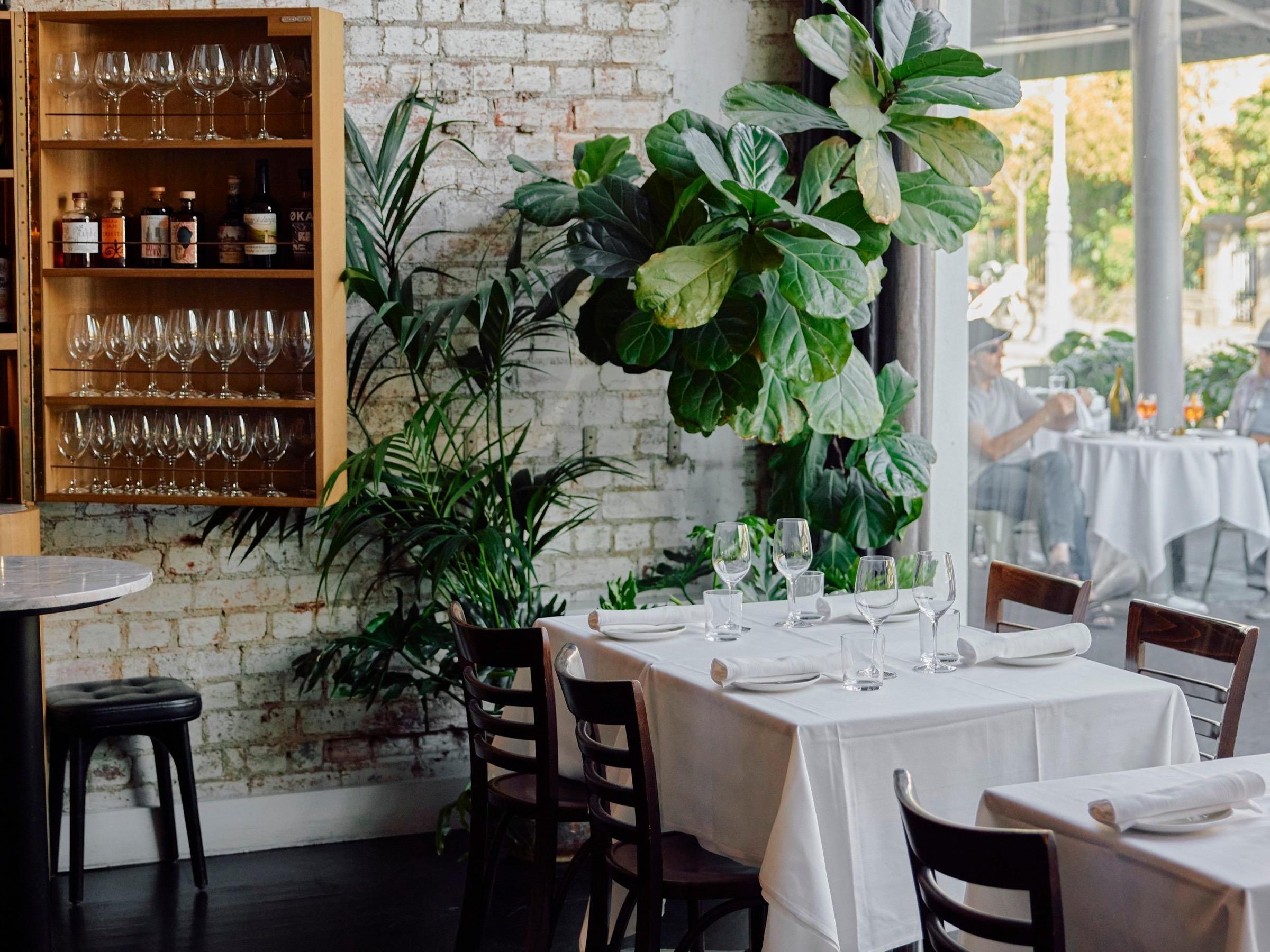 A bright dining room with white-clothed tables, shelves of glasses and potted plants
