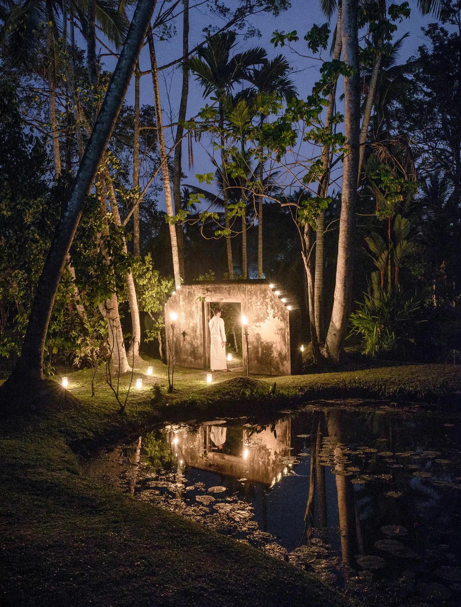 A small stone pavilion lit from within and surrounded by palm trees at night, its reflection visible in a lily pond.