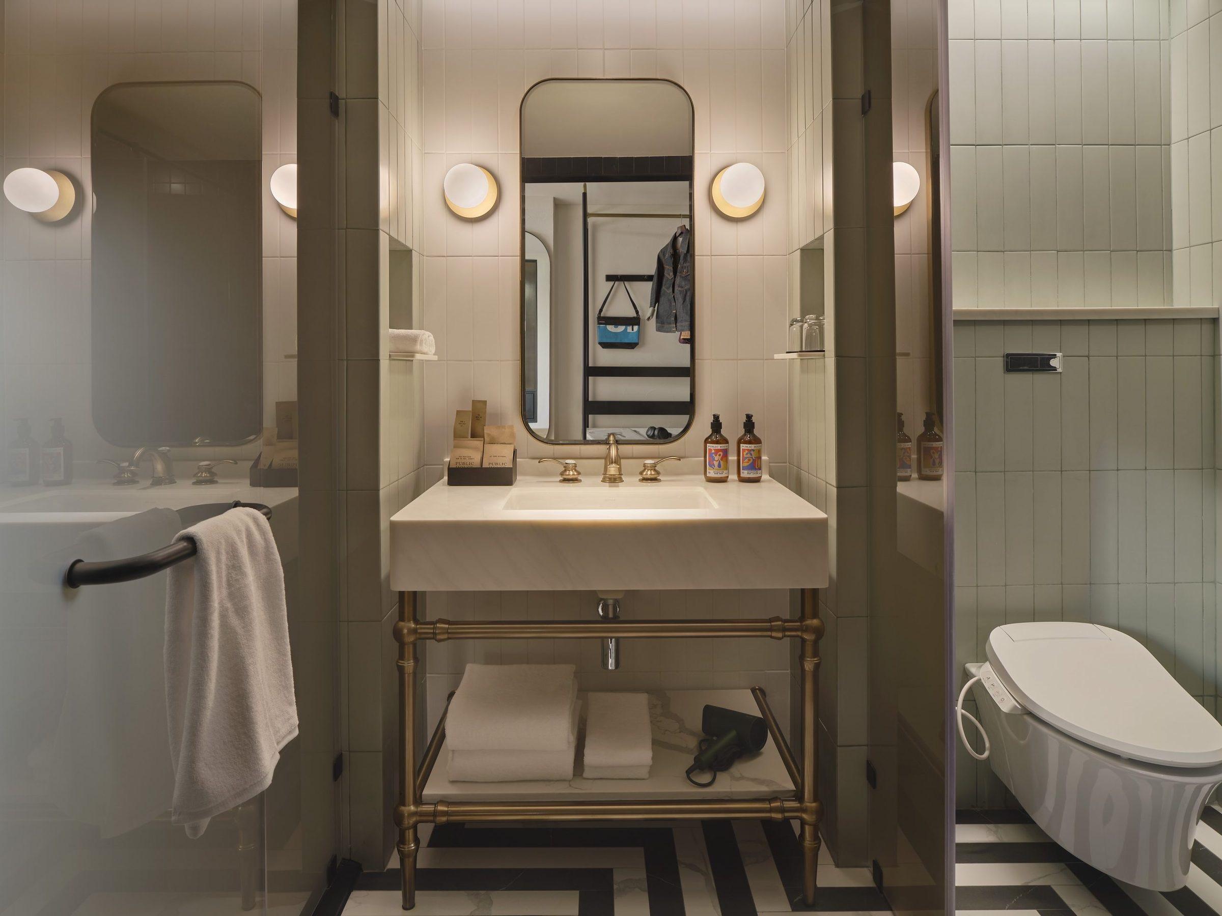 A bathroom with a marble topped vanity below a round mirror and globe wall lights, a freestanding bathtub to one side and a wall-hung toilet on a black and white chevron tiled floor.