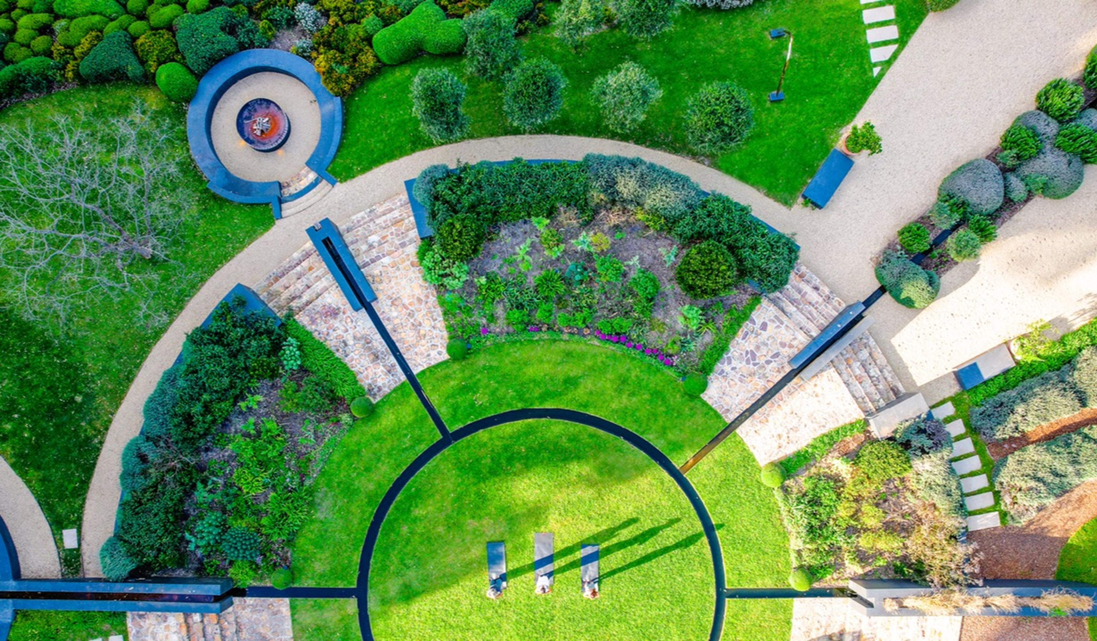 An aerial view straight down onto a circular lawn with three sun loungers and long shadows, bordered by curved stone paths, clipped hedges and a flower bed in bloom.