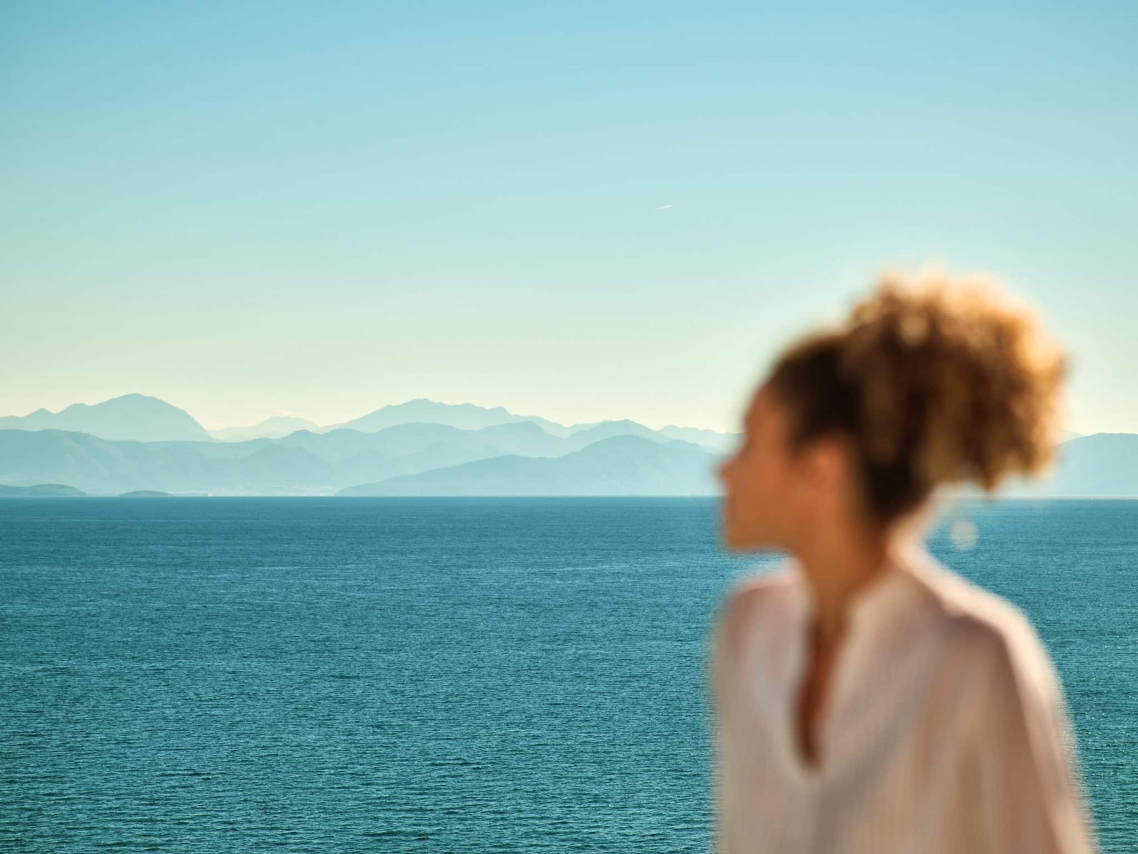 A woman in a white shirt, out of focus in the foreground, looking out over the sea toward a line of distant mountains.