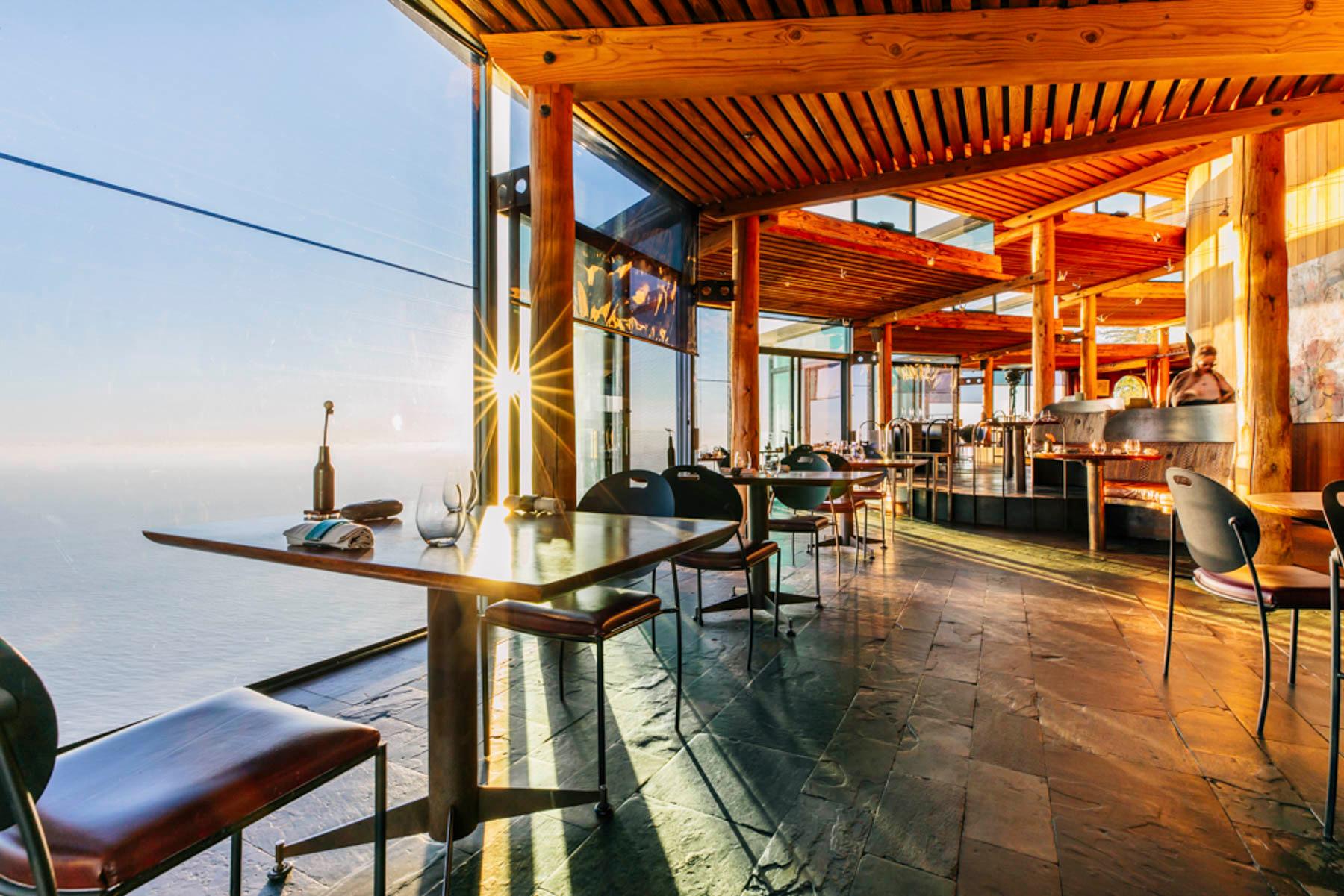 A dining room walled in glass on a cliff, tables set by the windows and the Pacific far below.