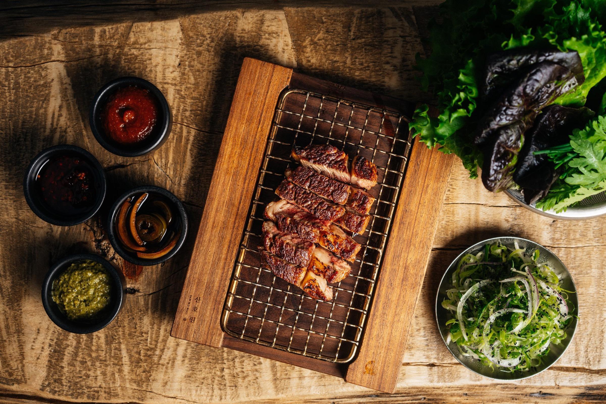 Grilled short rib on a charcoal grate on a wooden board, with small dishes of sauce and greens around it