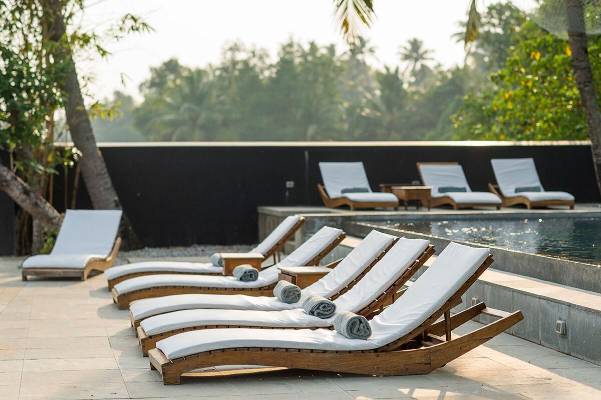 A row of wooden sun loungers with rolled towels beside an infinity pool, trees lit by early morning light beyond.