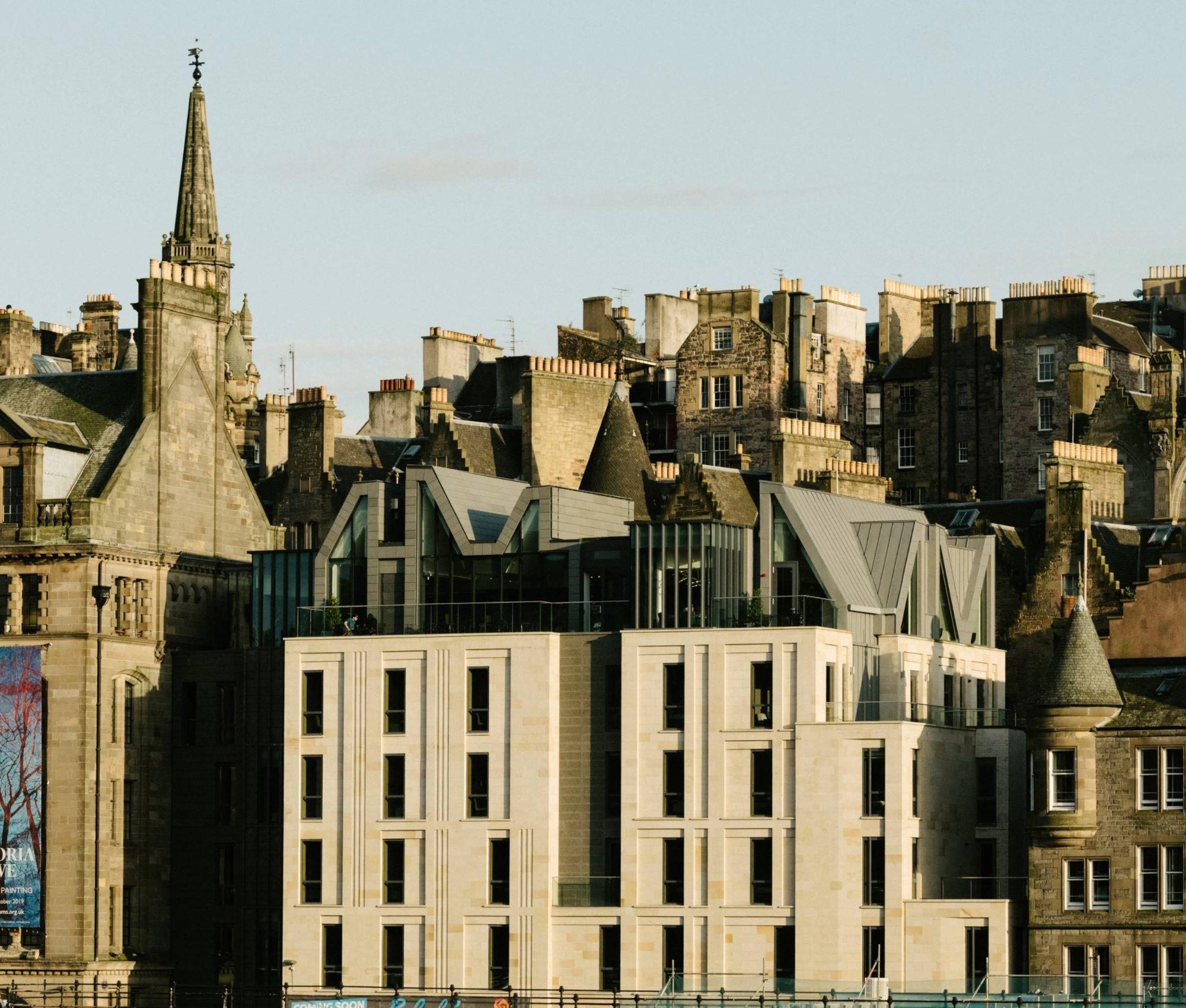 A row of Edinburgh buildings at dusk, including a pale stone building with glass-topped gables, seen above older stone townhouses and a church spire.