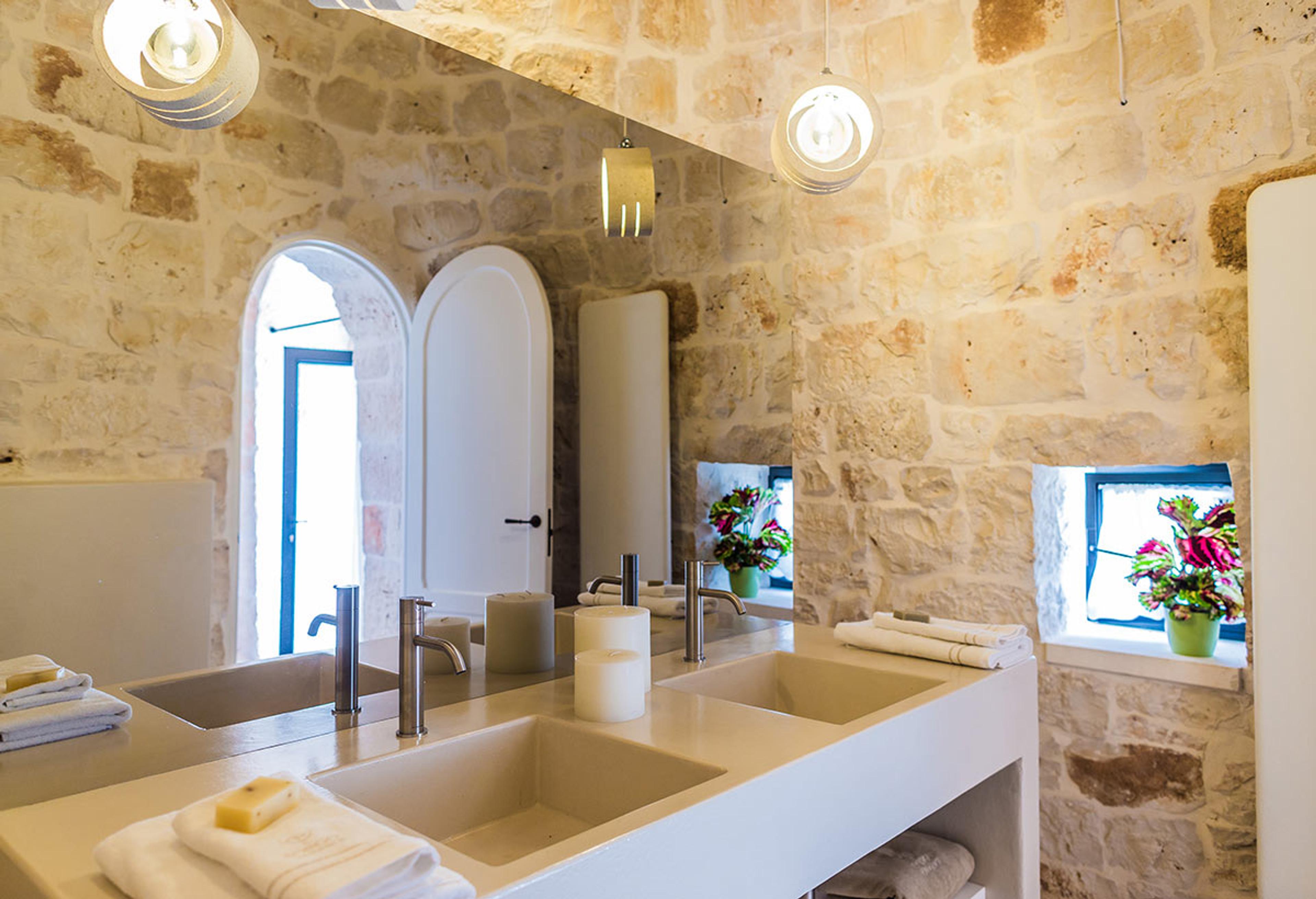 A stone bathroom with a double vanity, two basins, hanging pendant lights and a window filled with flowers.