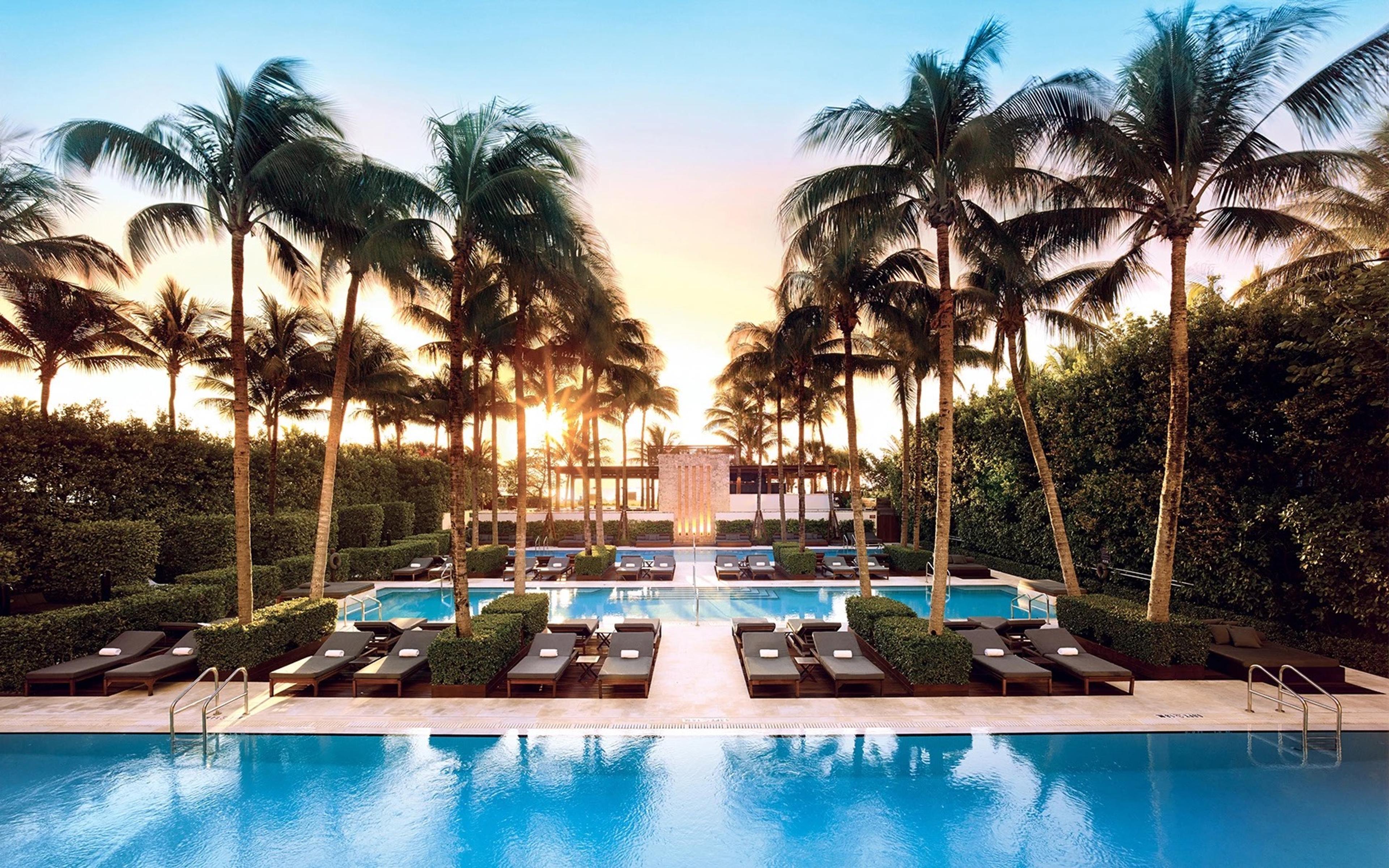 An avenue of palm trees frames two long rectangular pools and sun loungers, with the low-rise hotel and a golden sunset beyond.