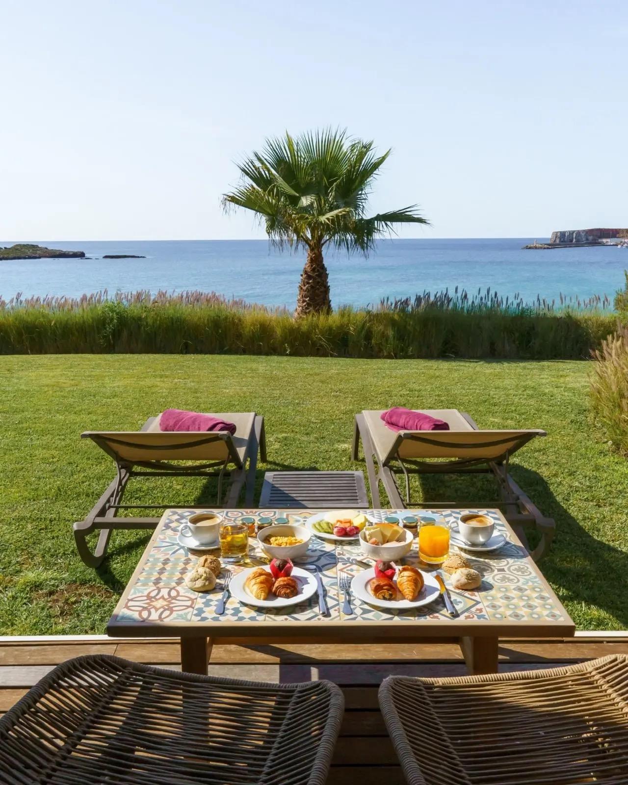 A breakfast table on a lawn with plates of croissants, fruit, coffee and orange juice, two loungers, a palm tree and the sea beyond.