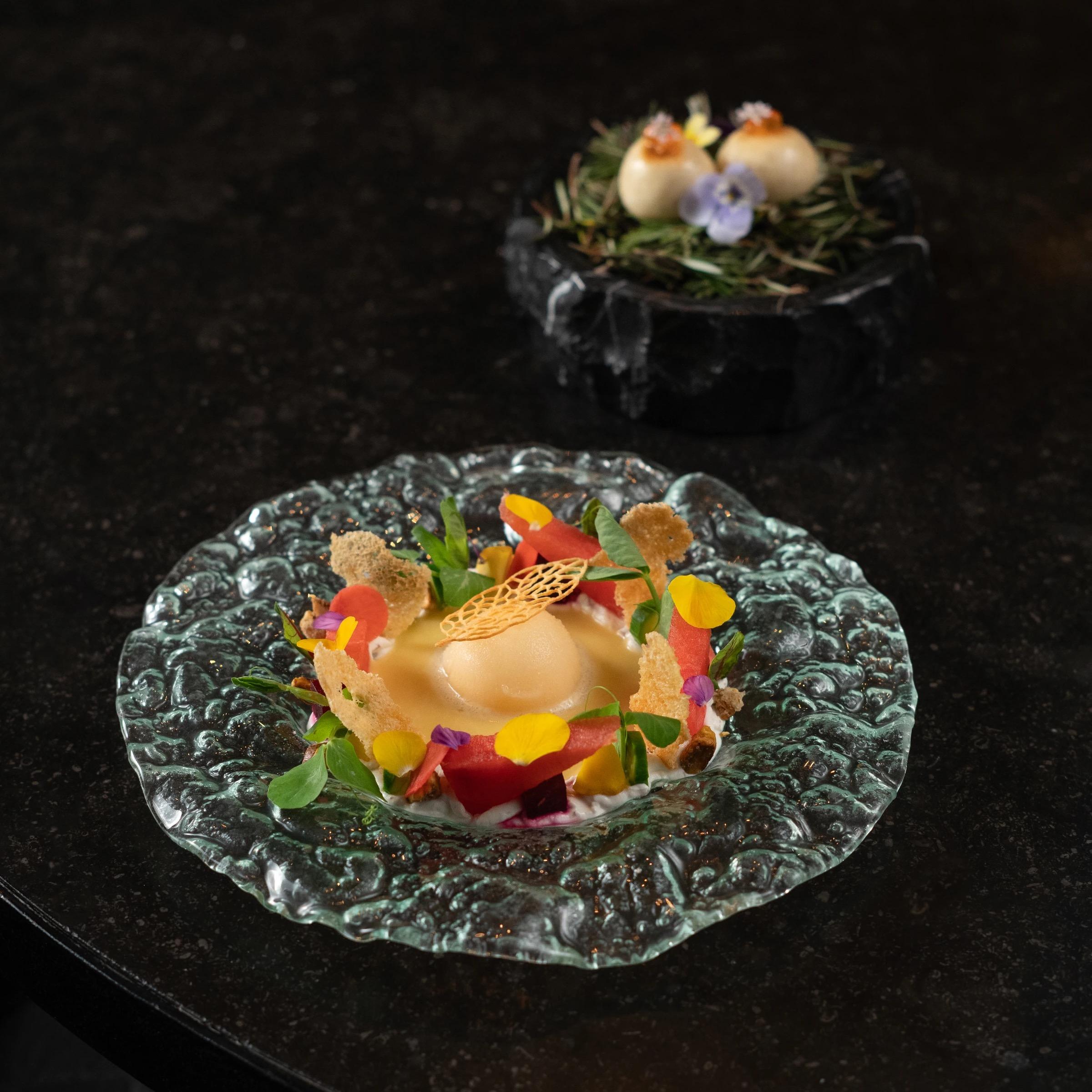 A plated dish of scallop and watermelon with herbs and edible flowers on a textured glass plate, with two filled pastries on a dark board behind.