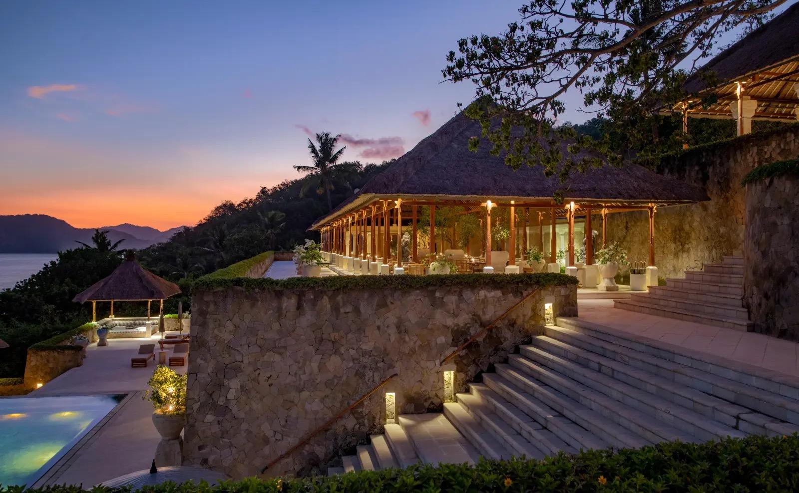 A stone terrace and steps lead up to an open-sided pavilion lit at dusk, with the sea visible beyond the hillside.