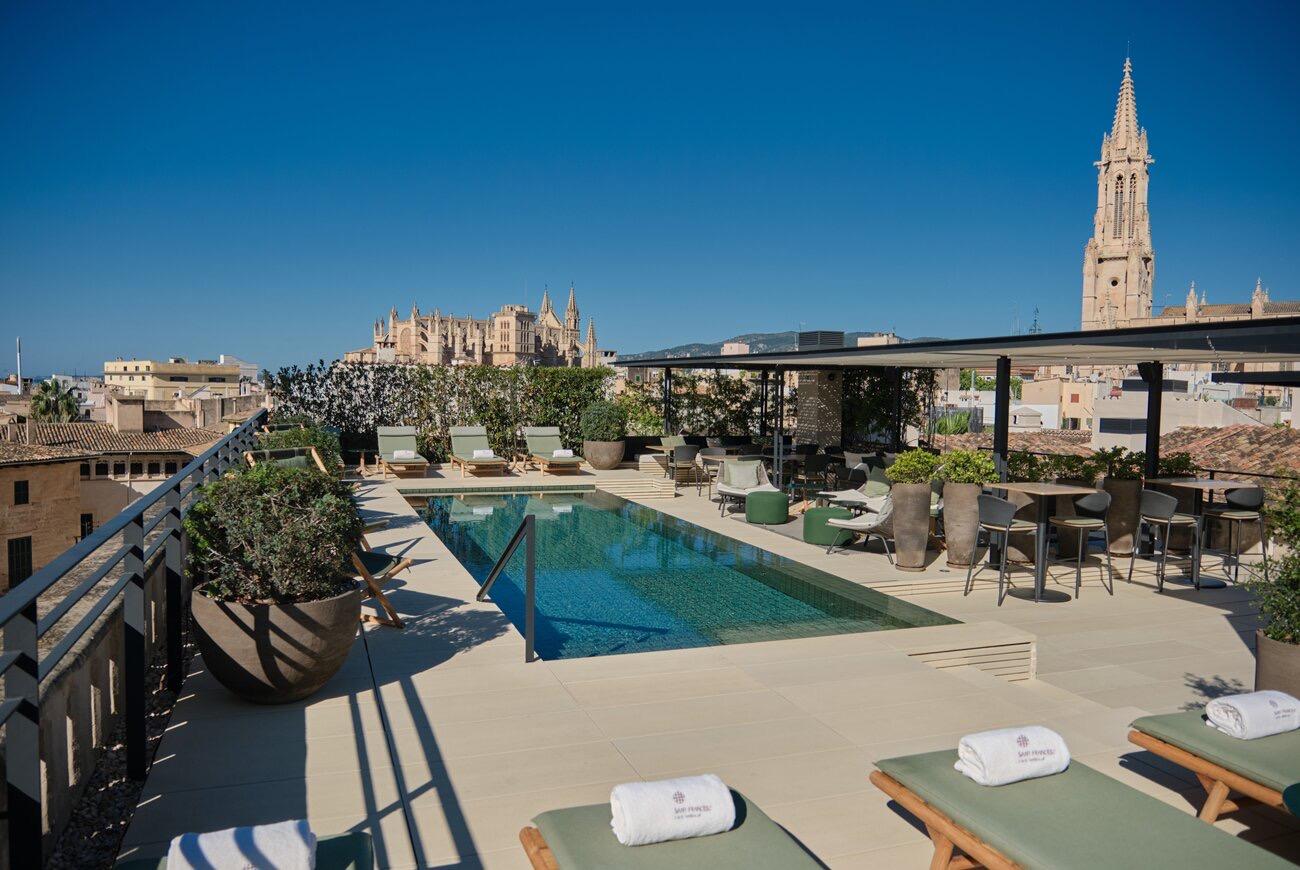 A rooftop terrace with a pool, a pergola-shaded seating area beyond it, cream loungers with rolled towels in the foreground, and a tall stone church tower rising over the city behind.