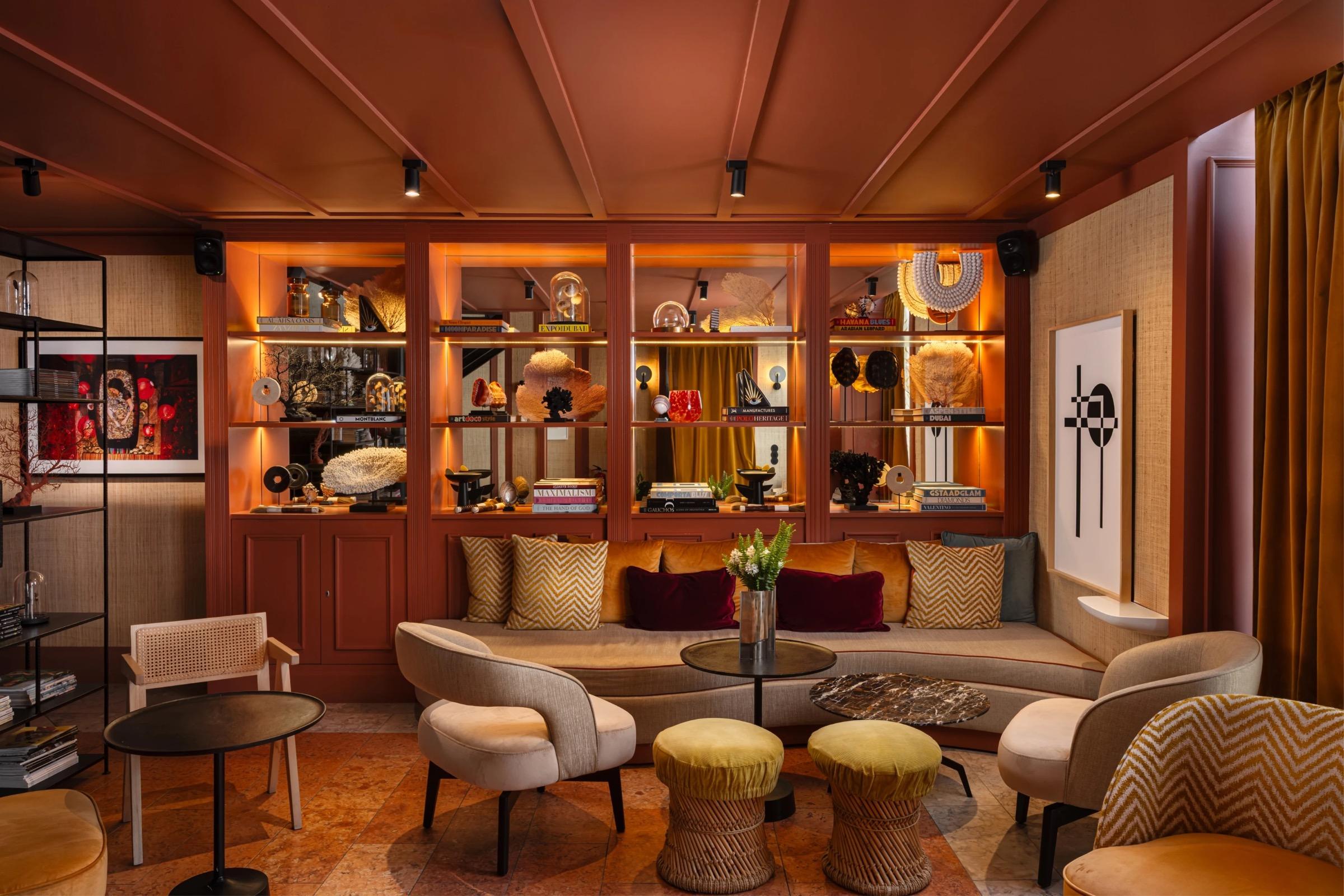 A lounge with burnt-orange walls and ceiling, lit shelves of ornaments and books behind a curved sofa with yellow and maroon cushions, and two velvet stools on a cork floor.
