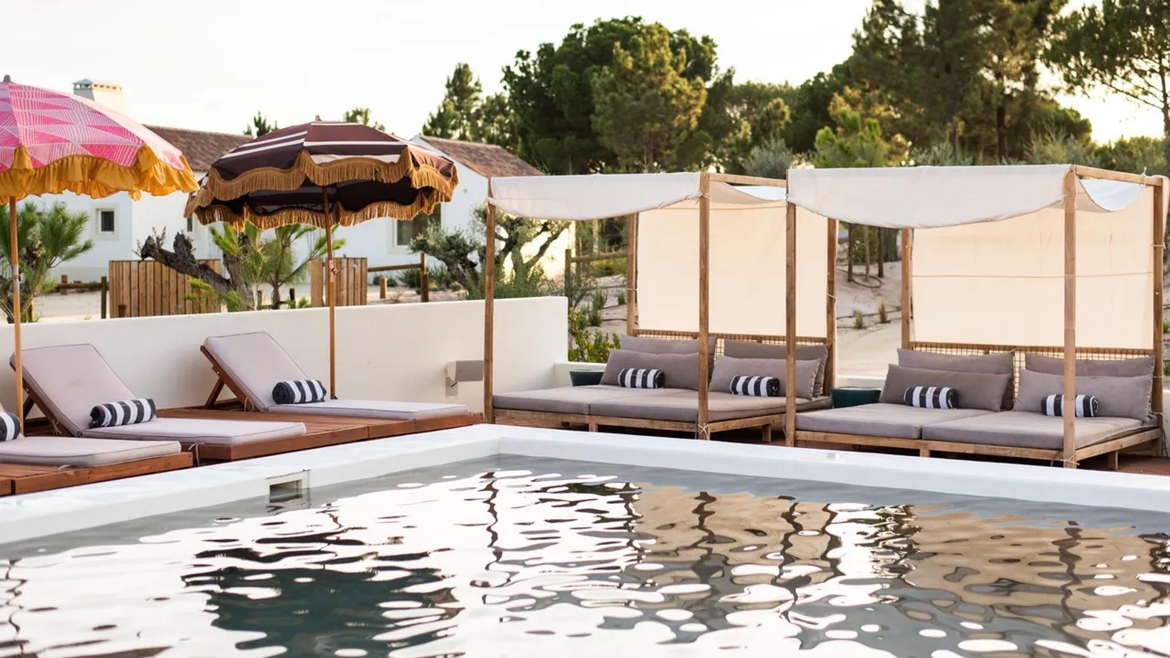 a swimming pool with loungers and patterned umbrellas on one side and canopied daybeds on the other, reflections of the sky in the water