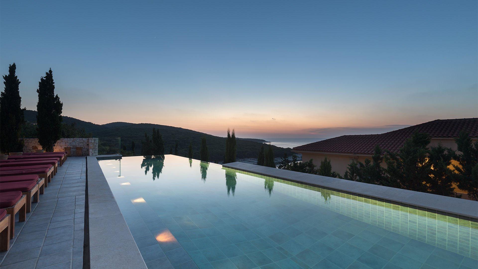 An infinity pool at dusk, red loungers along one edge, wooded hills and the sea beyond under a darkening sky.