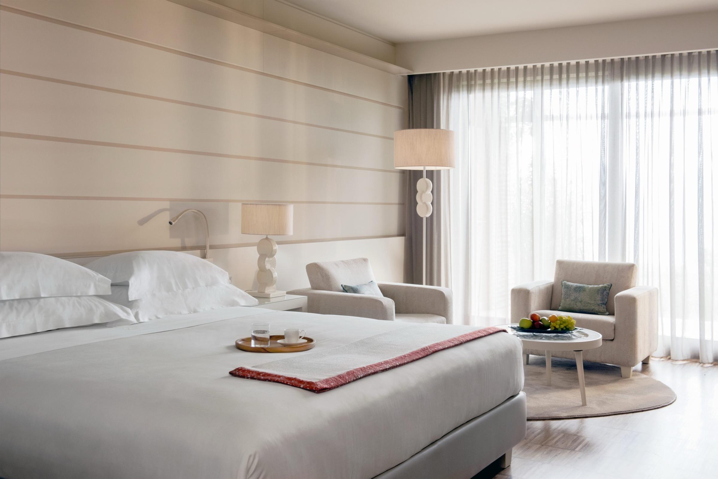A hotel bedroom with a wide bed, a seating area with two armchairs and a bowl of fruit, and full length windows along one wall.