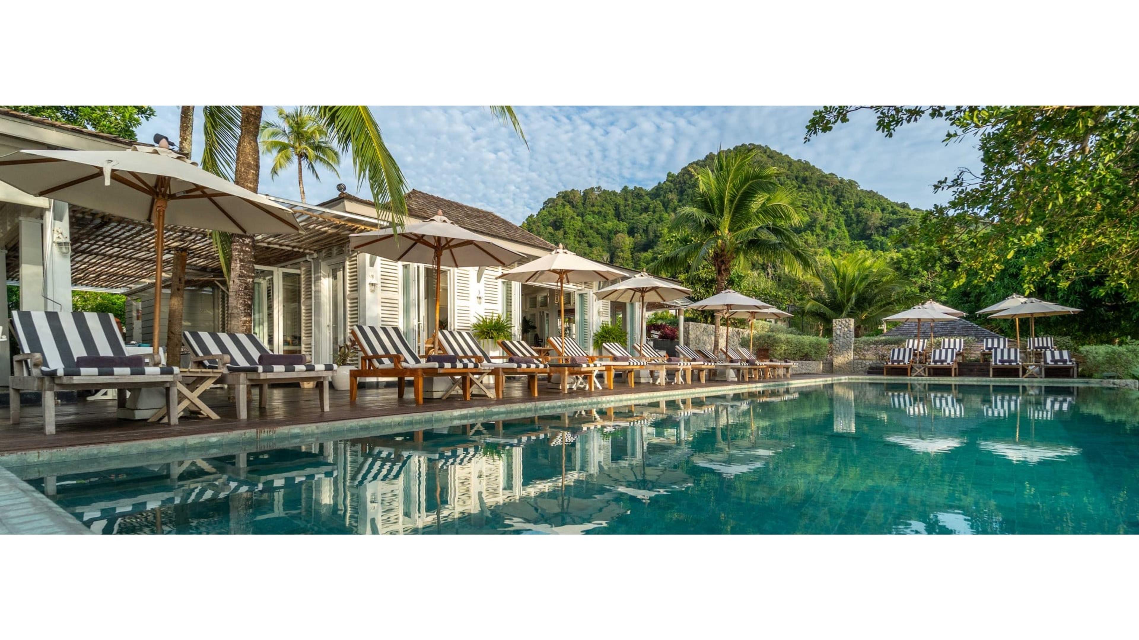 A pool running alongside a row of white shuttered buildings with striped loungers and umbrellas, backed by a forested limestone mountain.