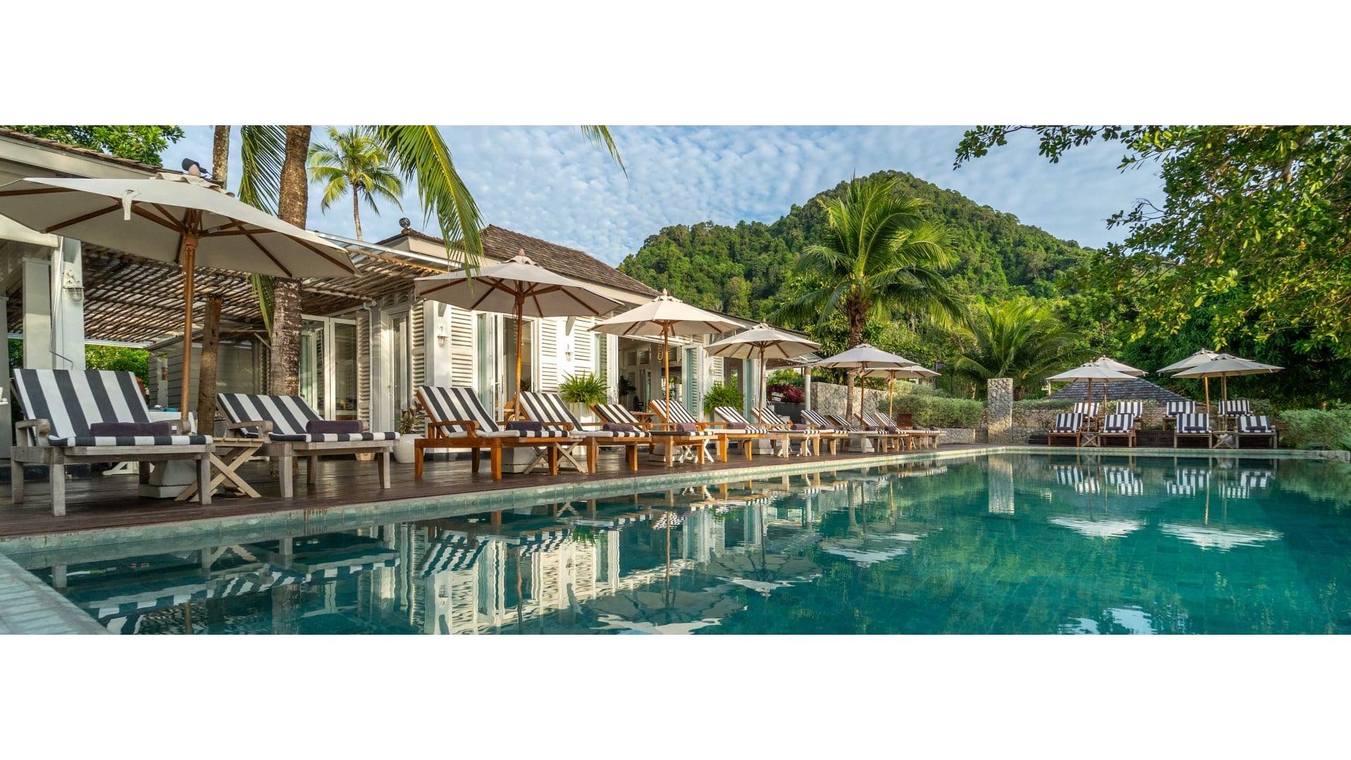 A pool running alongside a row of white shuttered buildings with striped loungers and umbrellas, backed by a forested limestone mountain.
