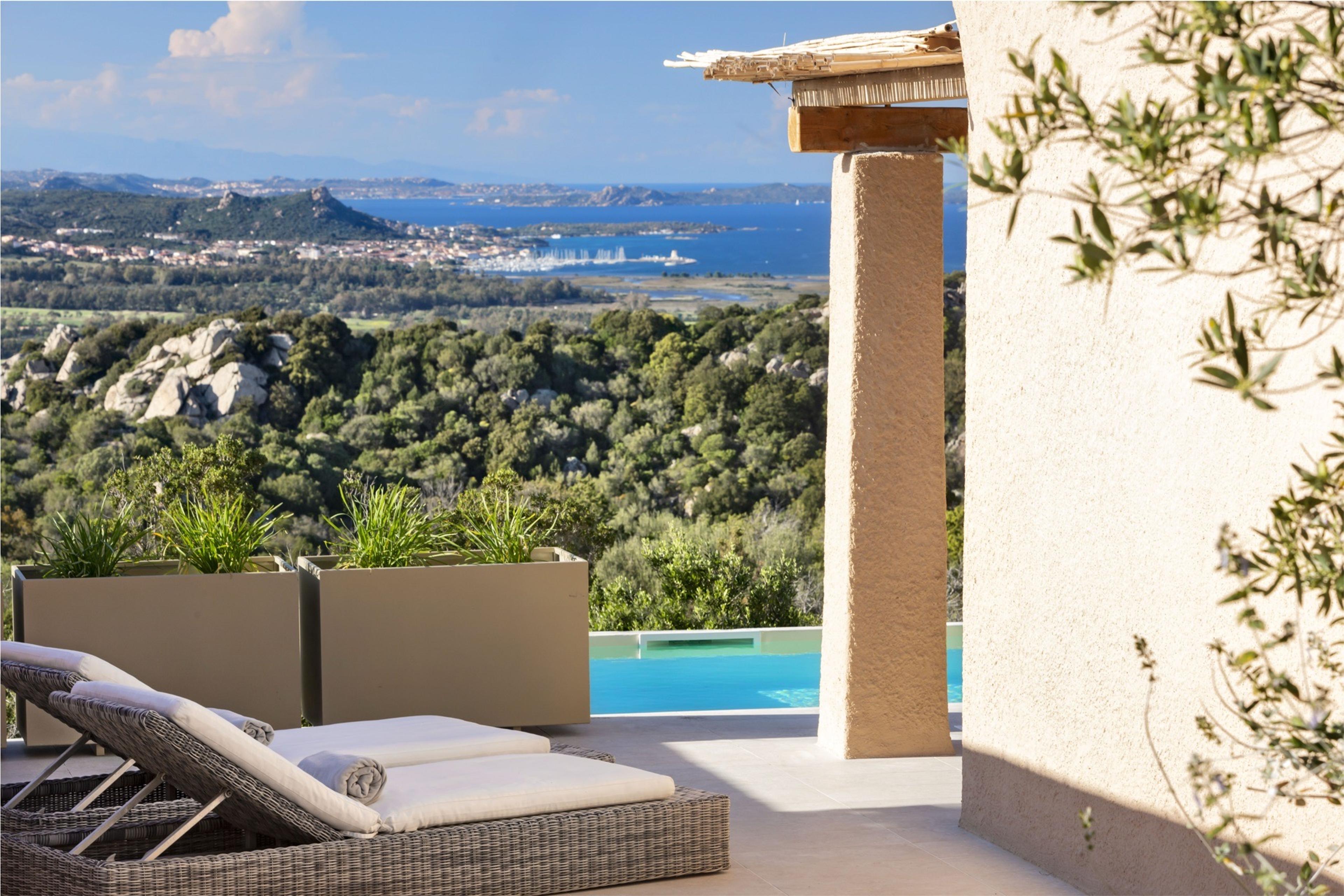 A terrace with wicker loungers beside an infinity pool, looking out over pine-covered hills to a coastal town, bay and islands in the distance.